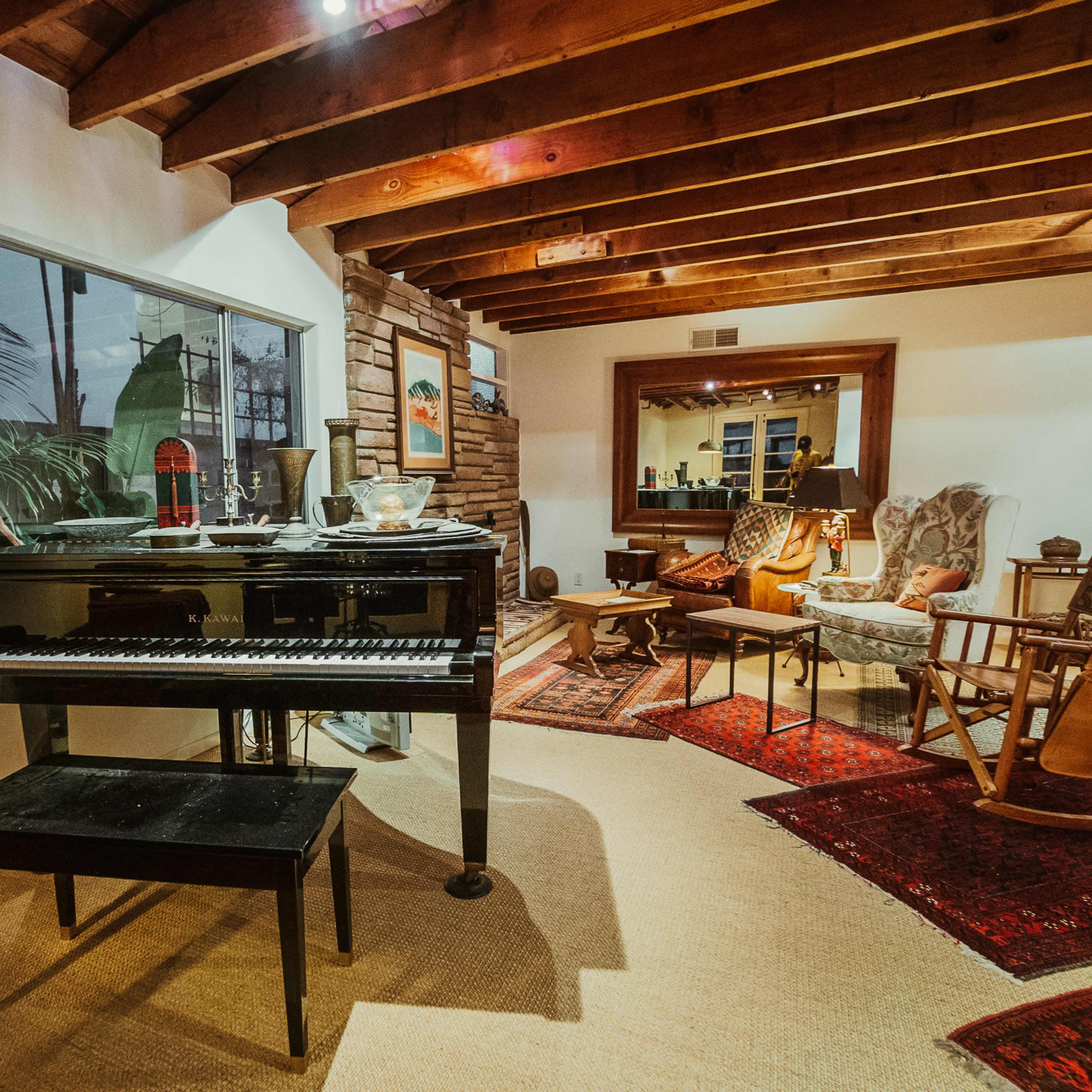 A black piano is positioned next to a window in a cozy living room featuring wooden beams, a rocking chair, and various decorative rugs.