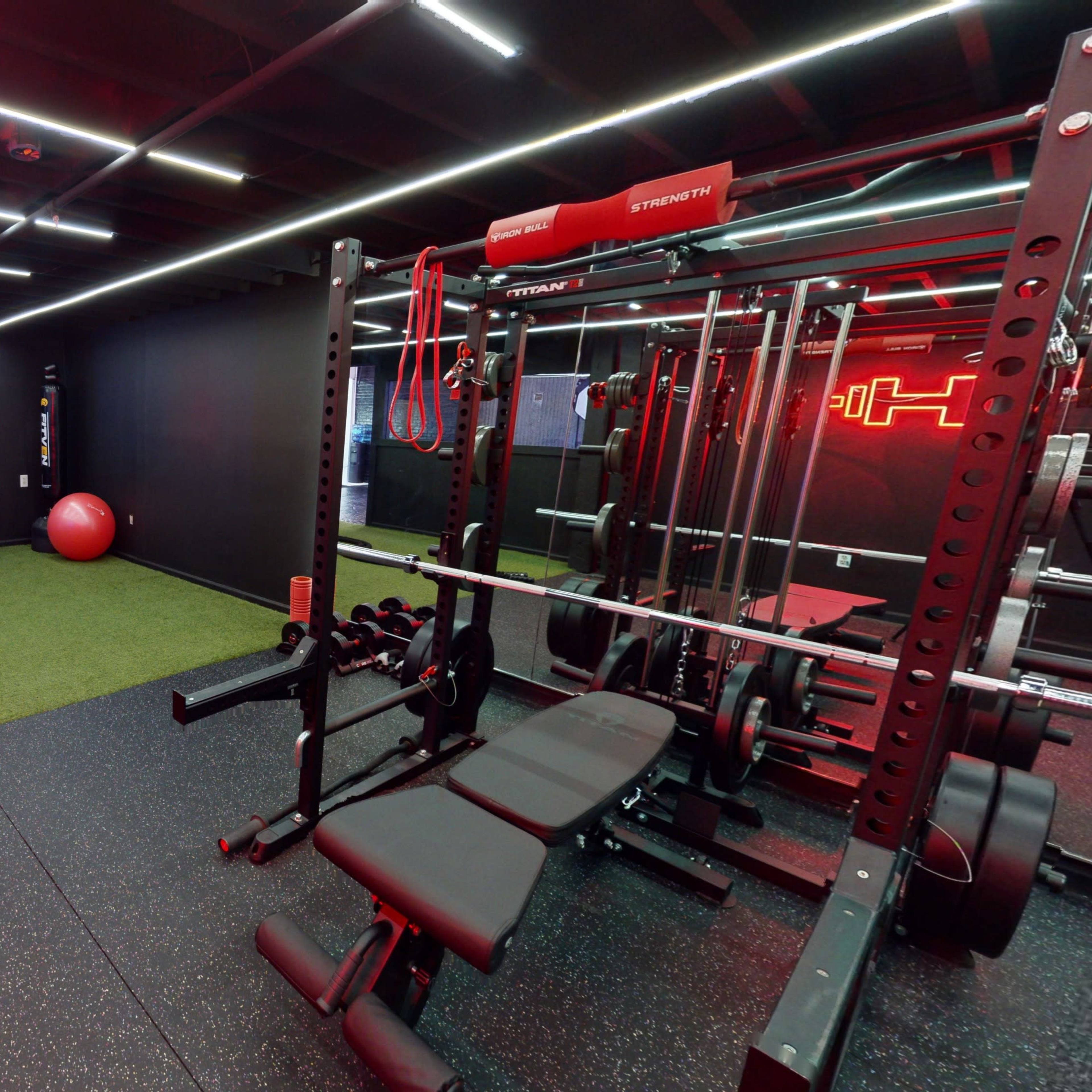 A modern gym interior featuring exercise equipment, a large red exercise ball, and artificial grass flooring under ambient lighting.