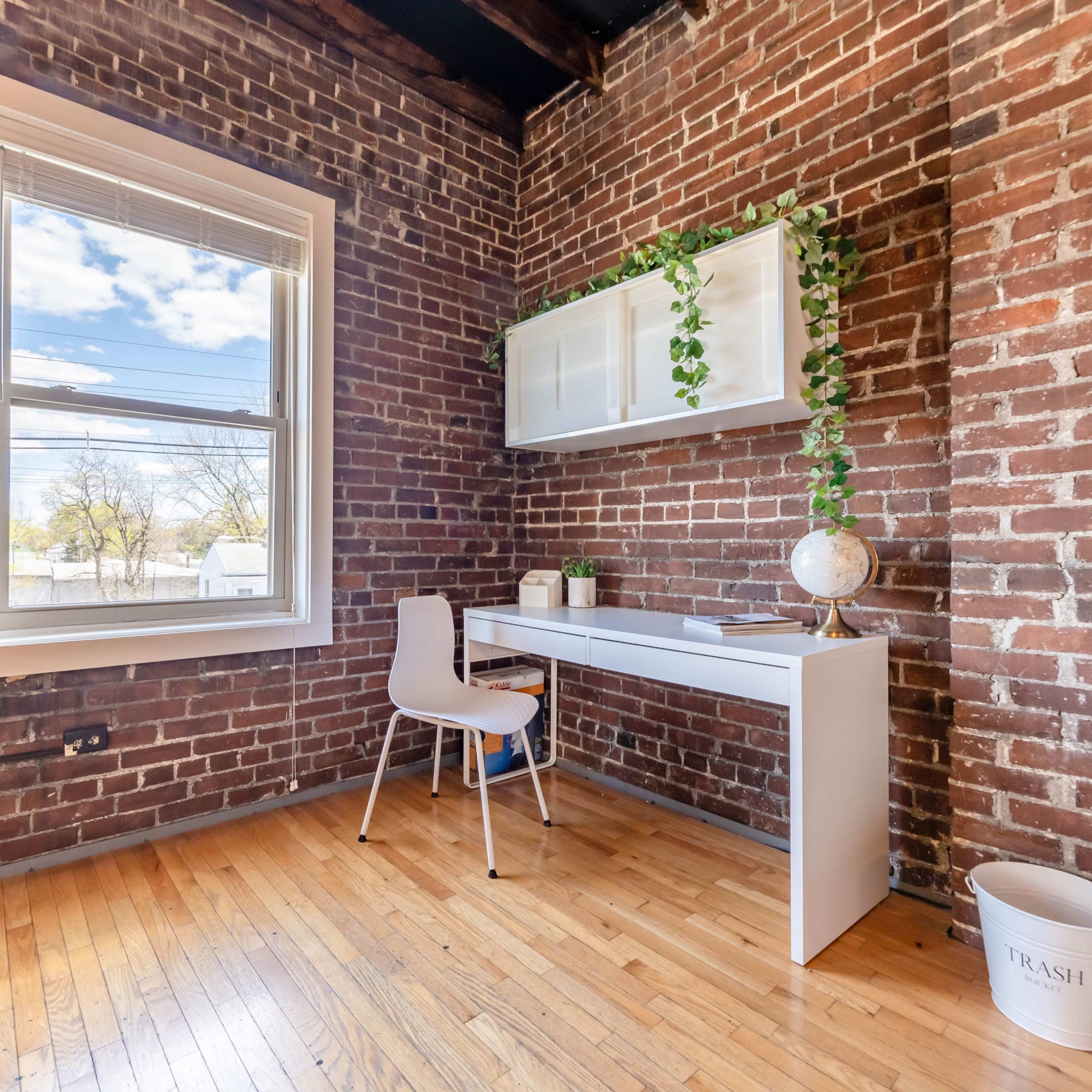 A small workspace with a white desk and chair against a brick wall, featuring a globe and a plant on the desk, with a window allowing natural light.