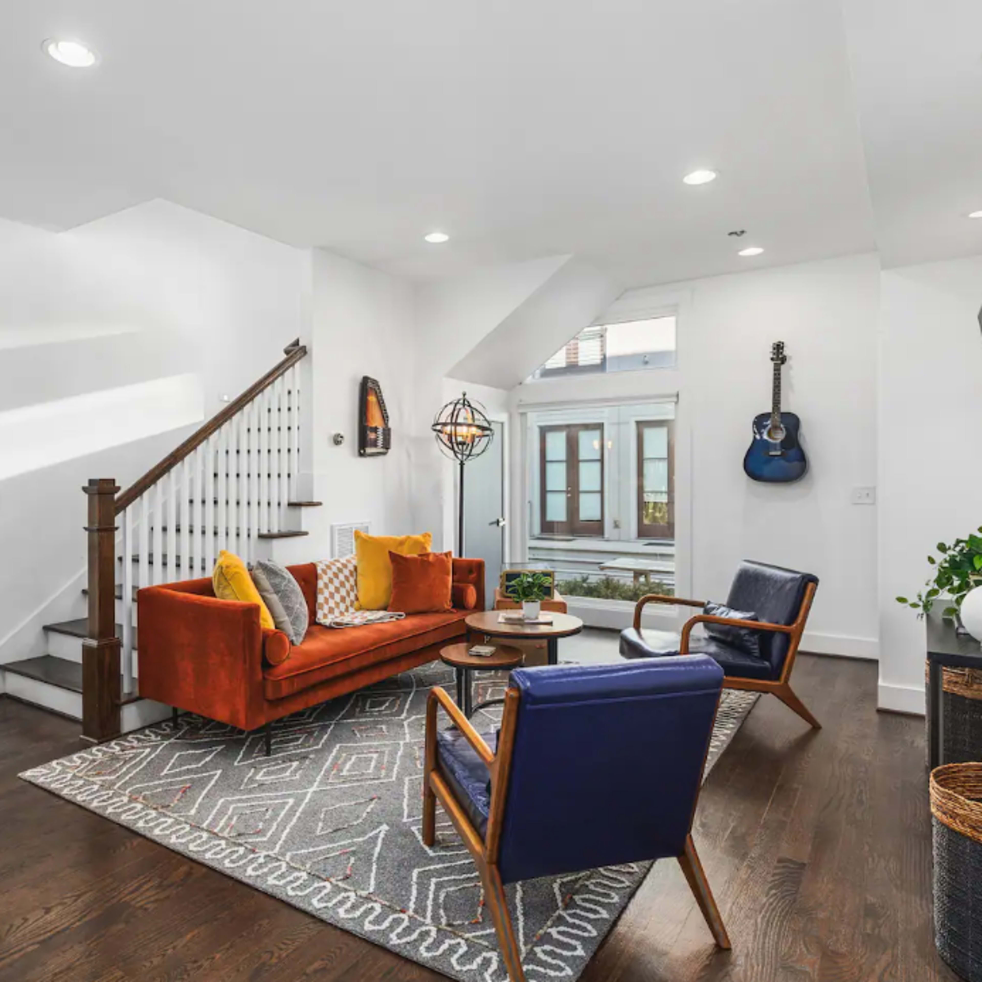 A modern living room featuring a red sofa, blue armchairs, a patterned area rug, and a staircase leading to an upper level.