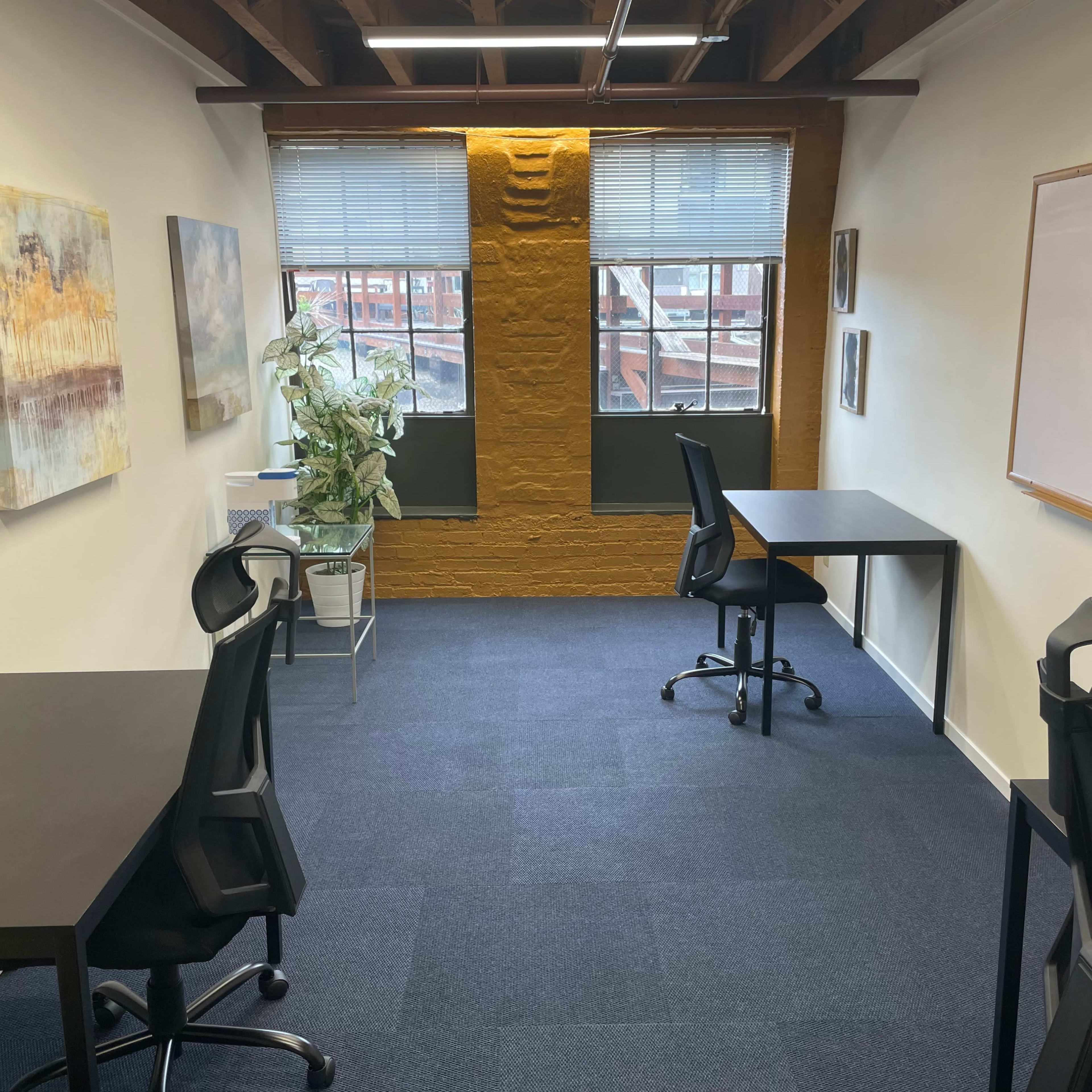 A modern office space with two black desks and chairs, large windows, and a plant, set against a textured yellow wall.