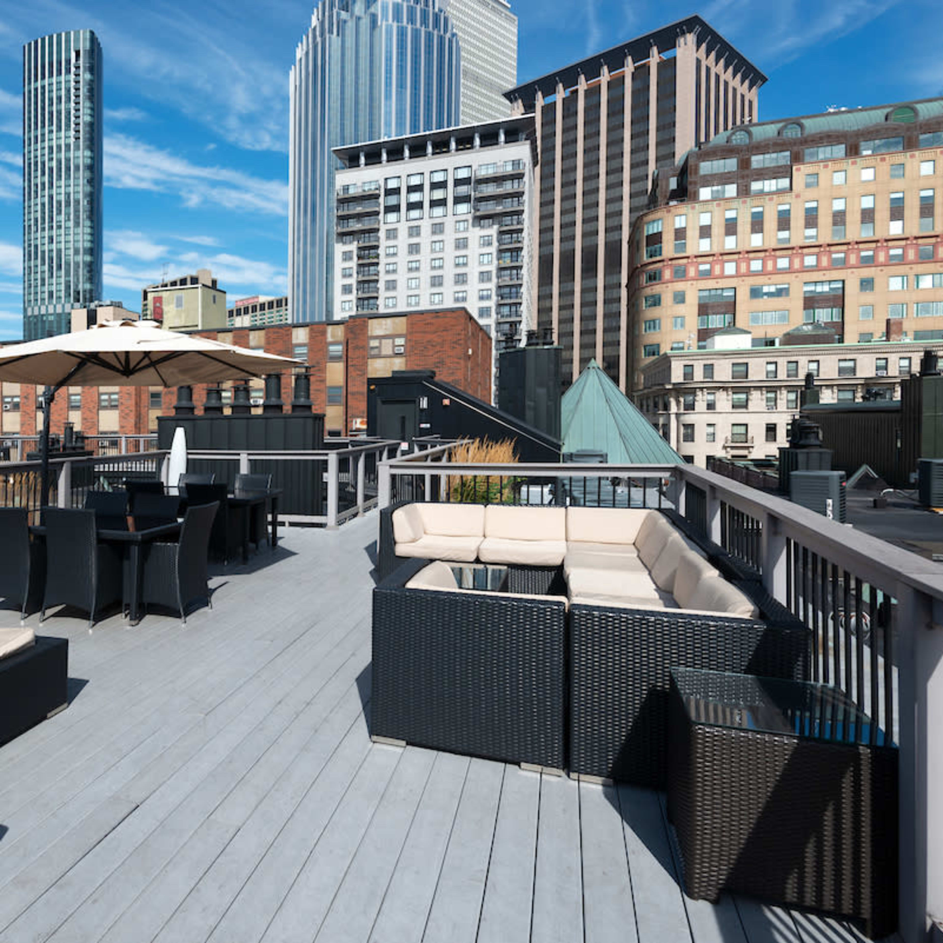 A modern rooftop patio features seating areas and an umbrella with a backdrop of tall city buildings.