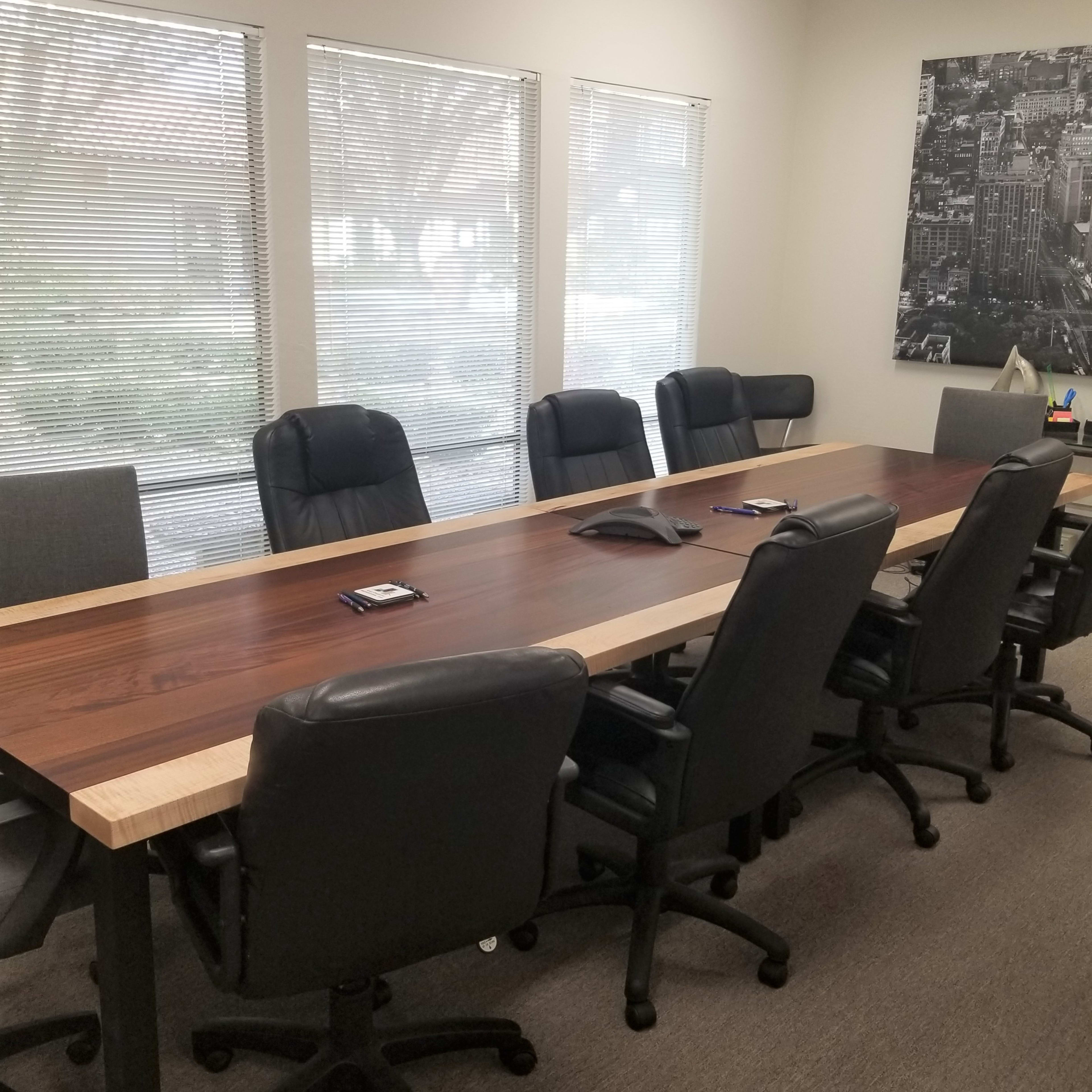 A conference room features a large wooden table surrounded by black rolling chairs, with windows letting in natural light.