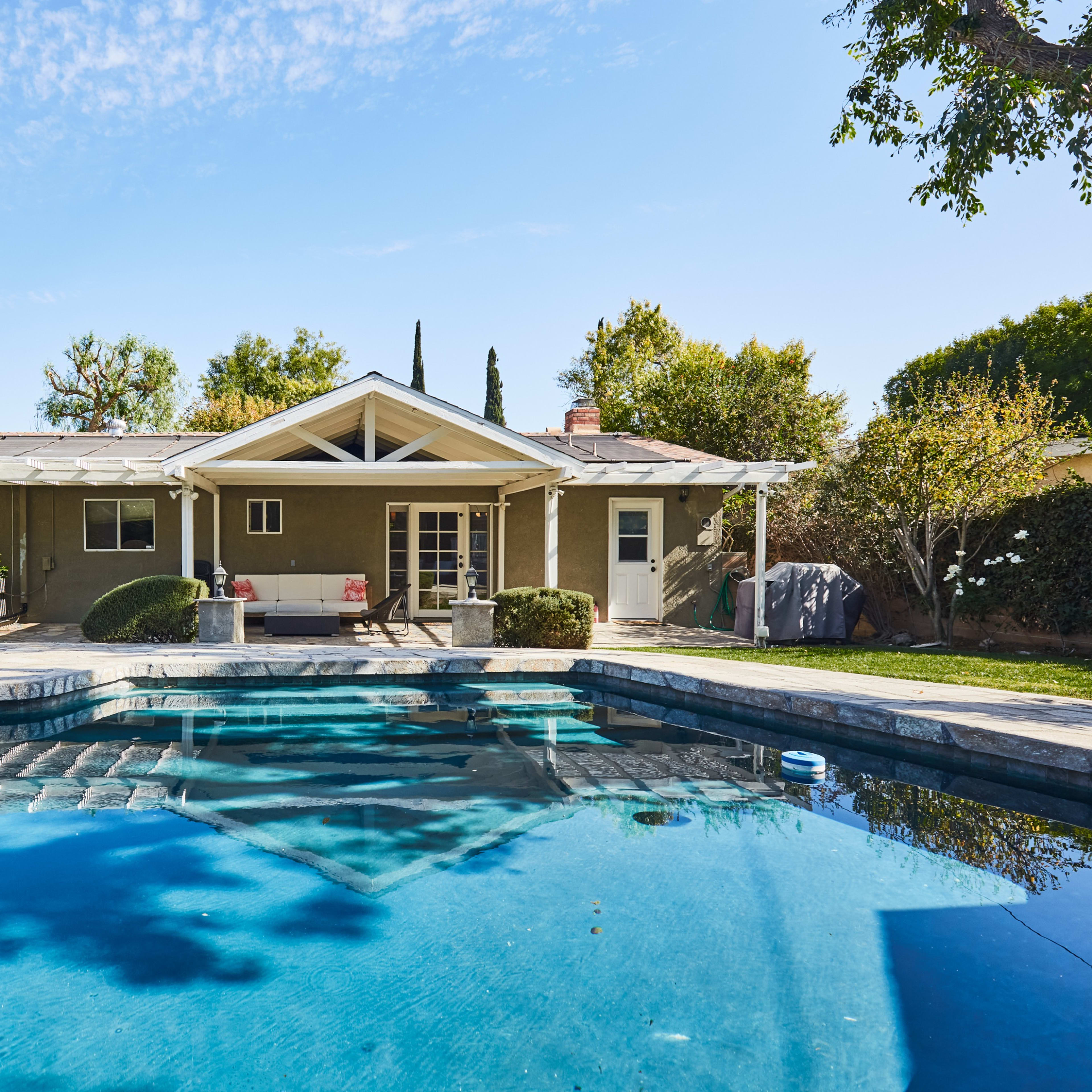 A backyard with a swimming pool in front of a single-story house, surrounded by trees and landscaped greenery.