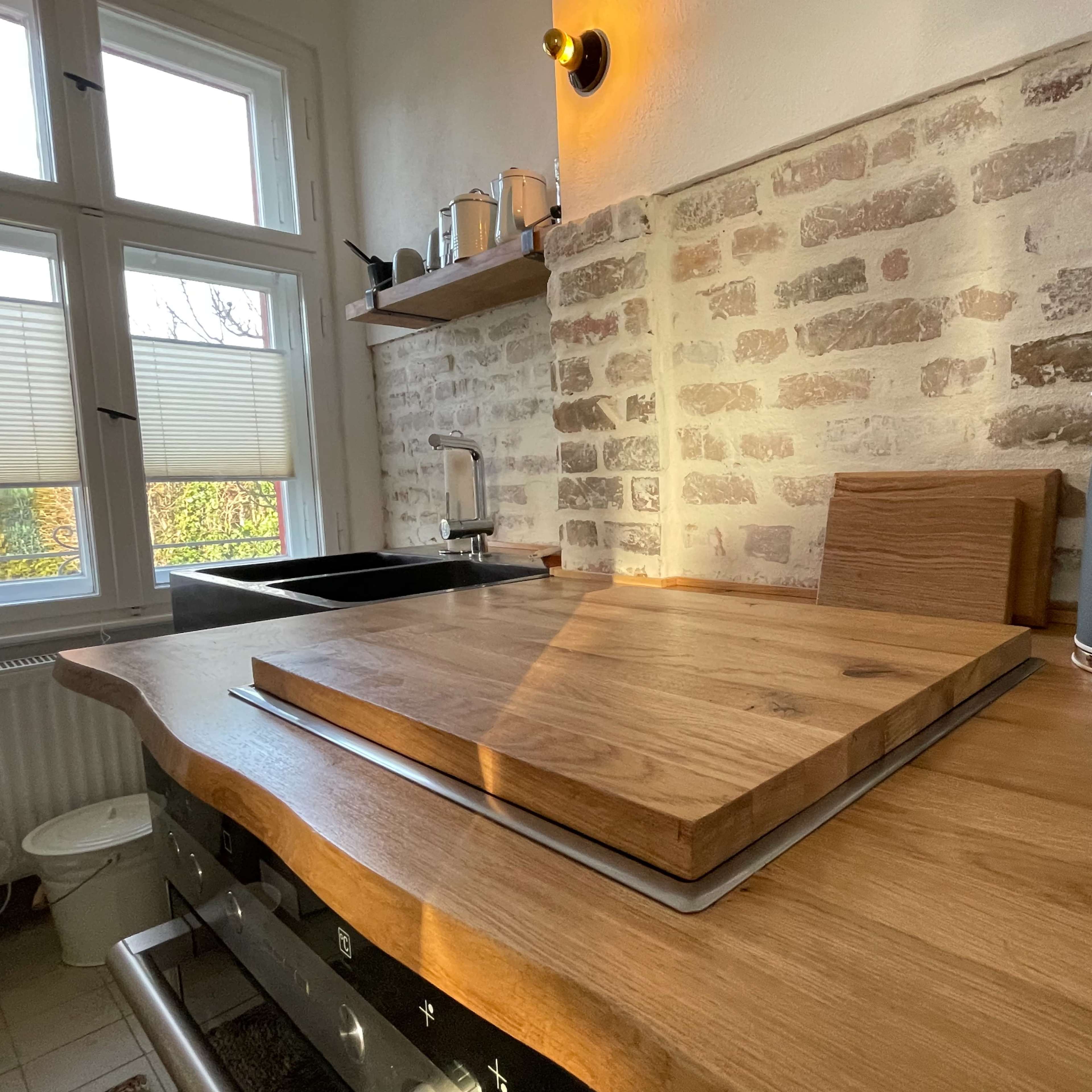A kitchen countertop made of wood with a chopping board, a kettle, and a sink under a window.