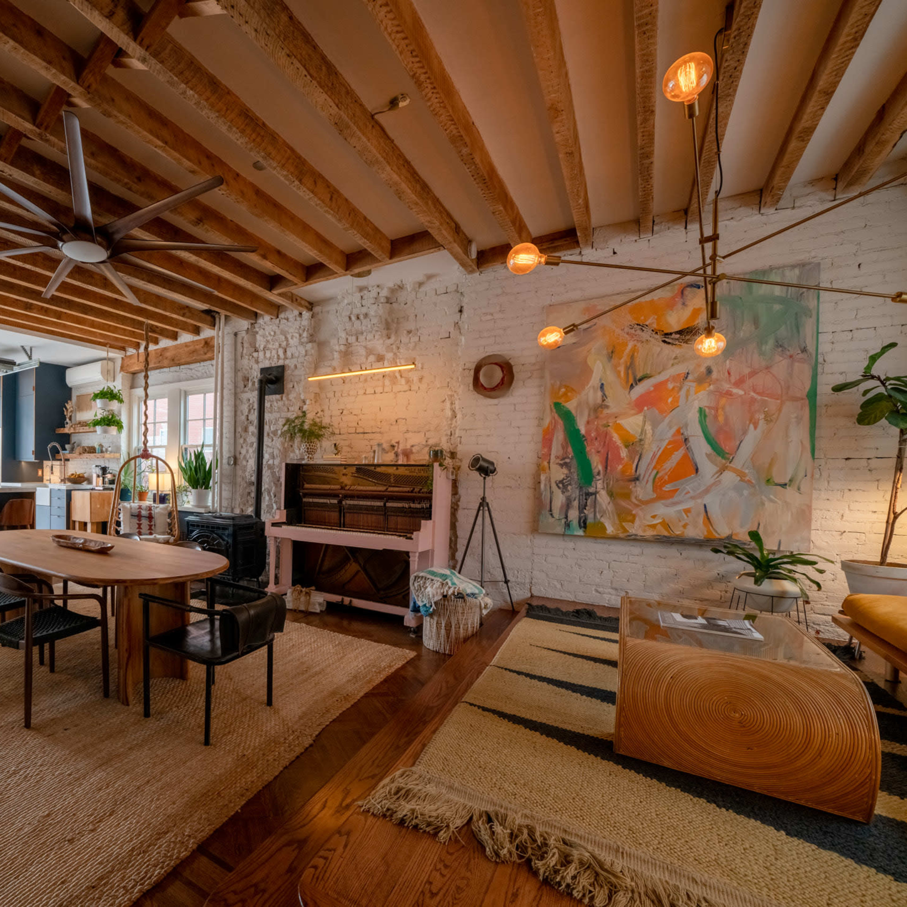 The scene features an open-concept living area with a kitchen, dining table, and vibrant artwork, highlighted by exposed wooden beams and ample natural light.