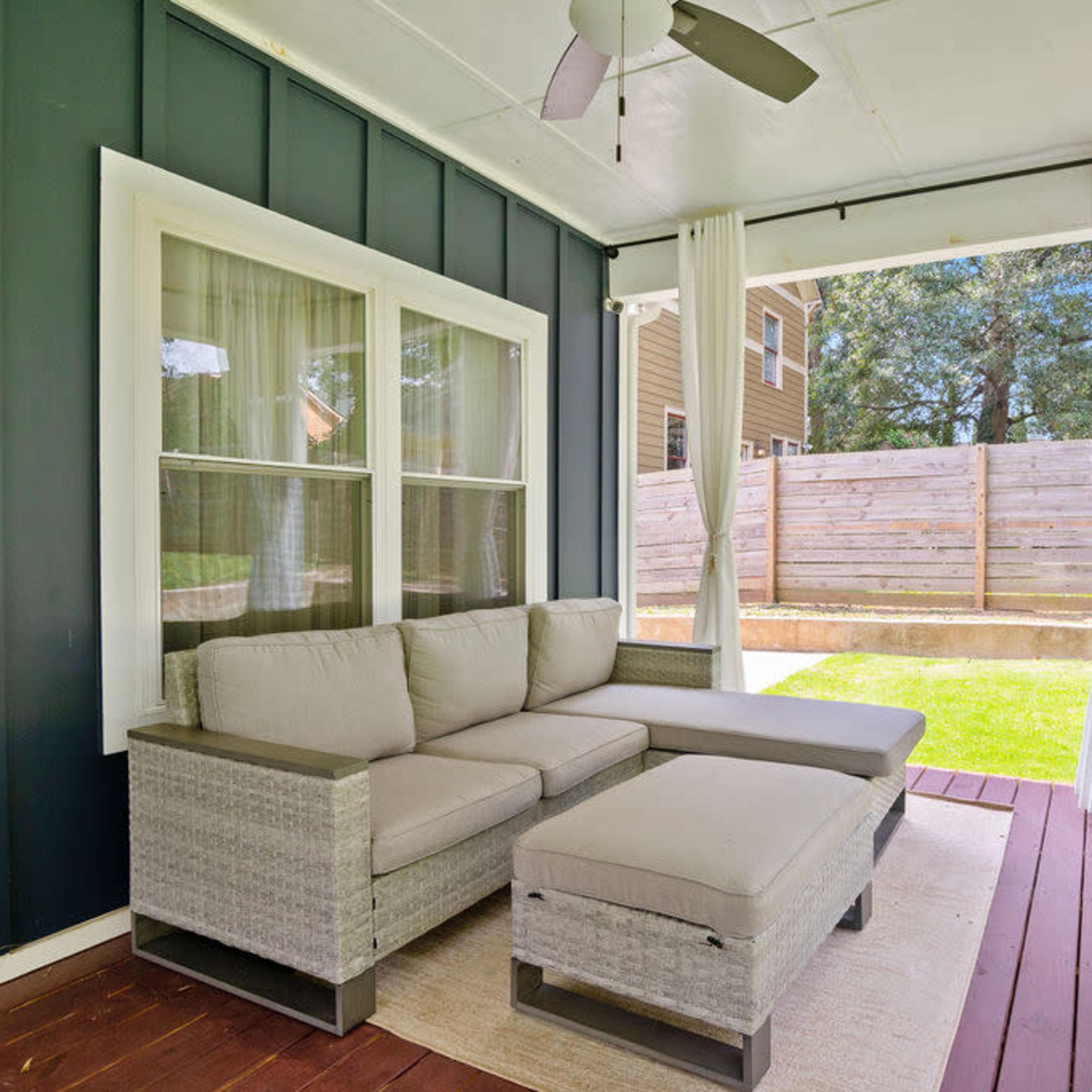 A covered porch features a gray sectional sofa and a matching ottoman, with sheer curtains flanking the windows.
