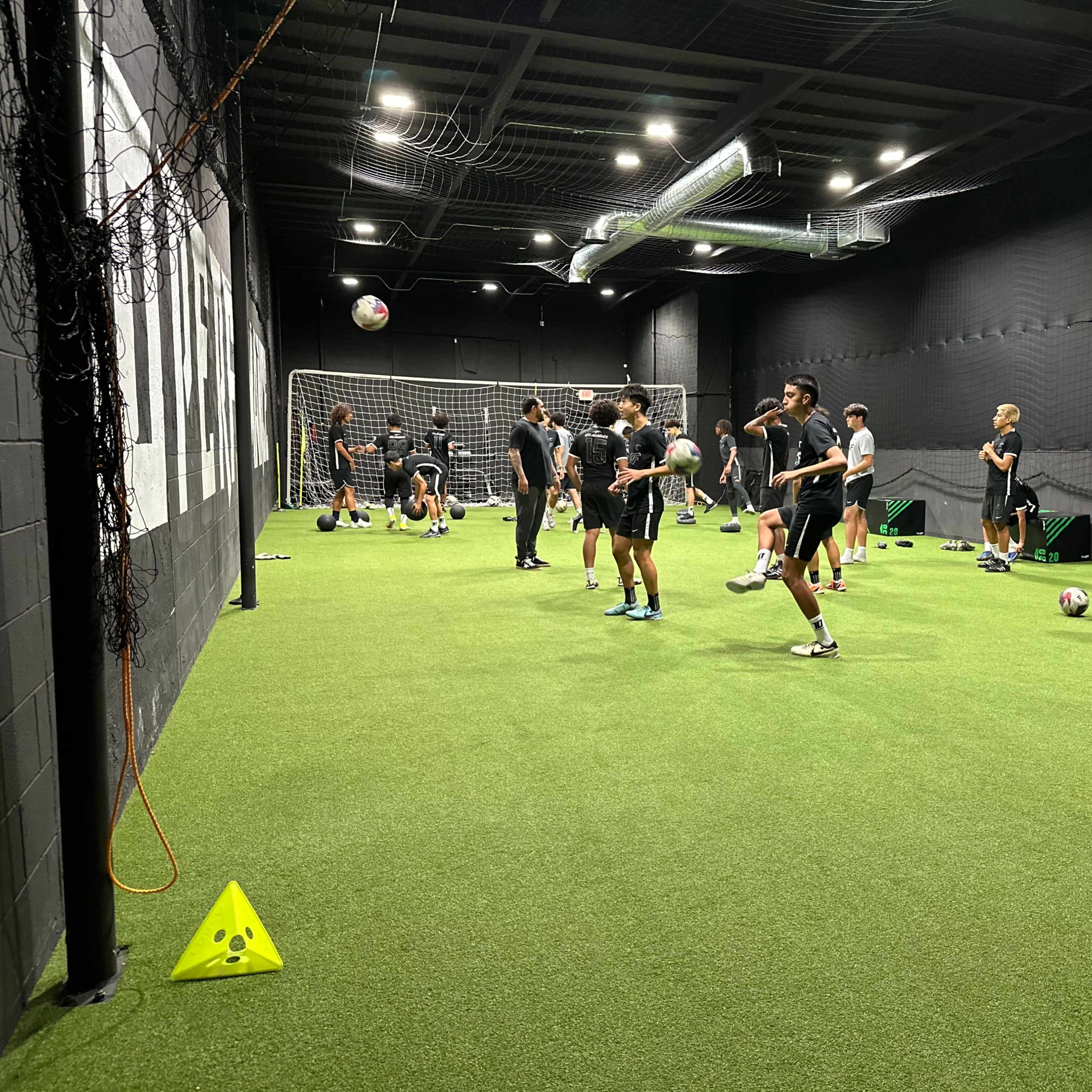 A group of athletes is practicing soccer drills in a carpeted indoor training facility.