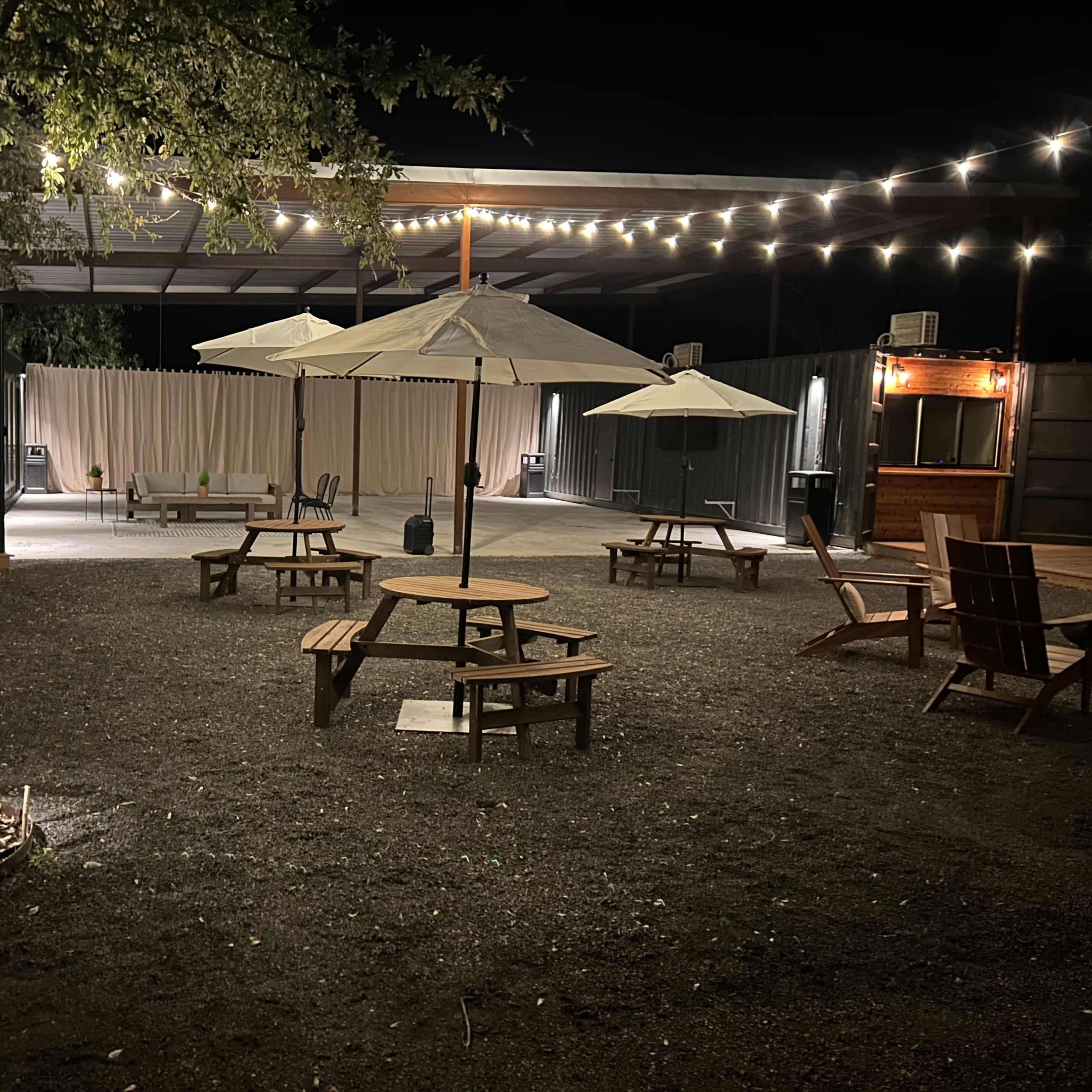 An outdoor gathering space at night, featuring several picnic tables and umbrellas, surrounded by string lights and a gravel ground.