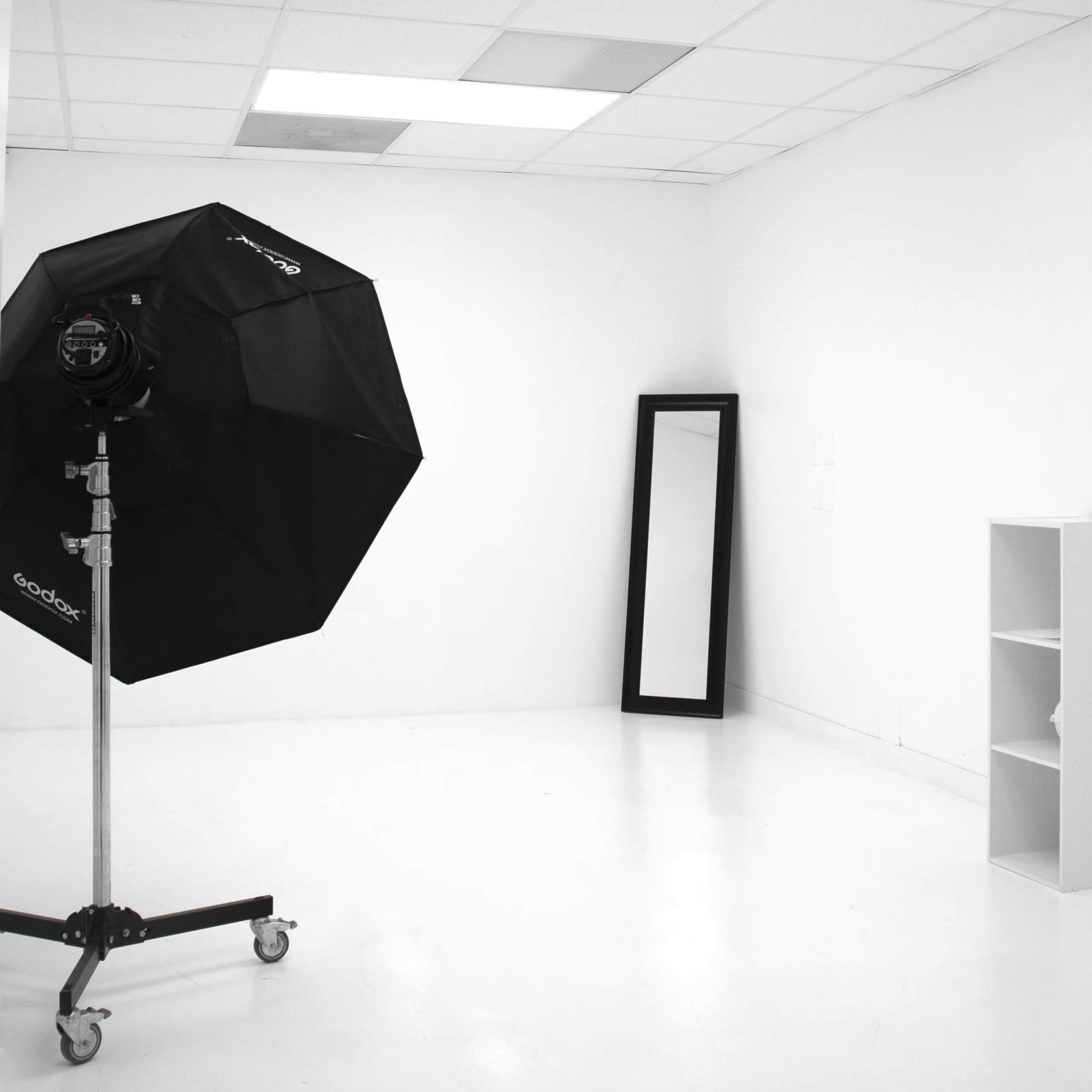 A minimalist white studio room with a large softbox light on a stand, a tall mirror, and a small shelving unit.