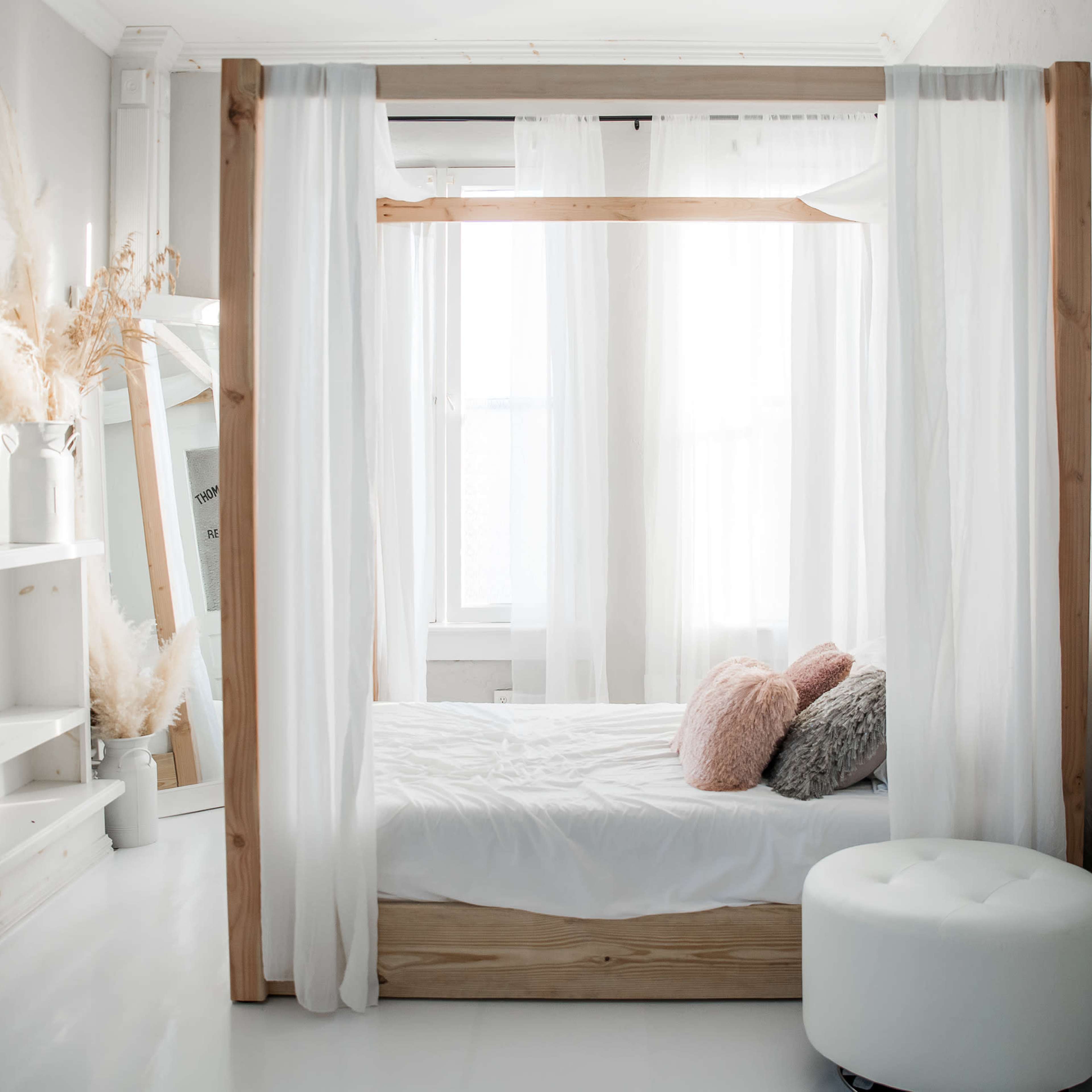 A four-poster bed with sheer curtains is positioned near a large window, accompanied by a round ottoman and decorative pillows.