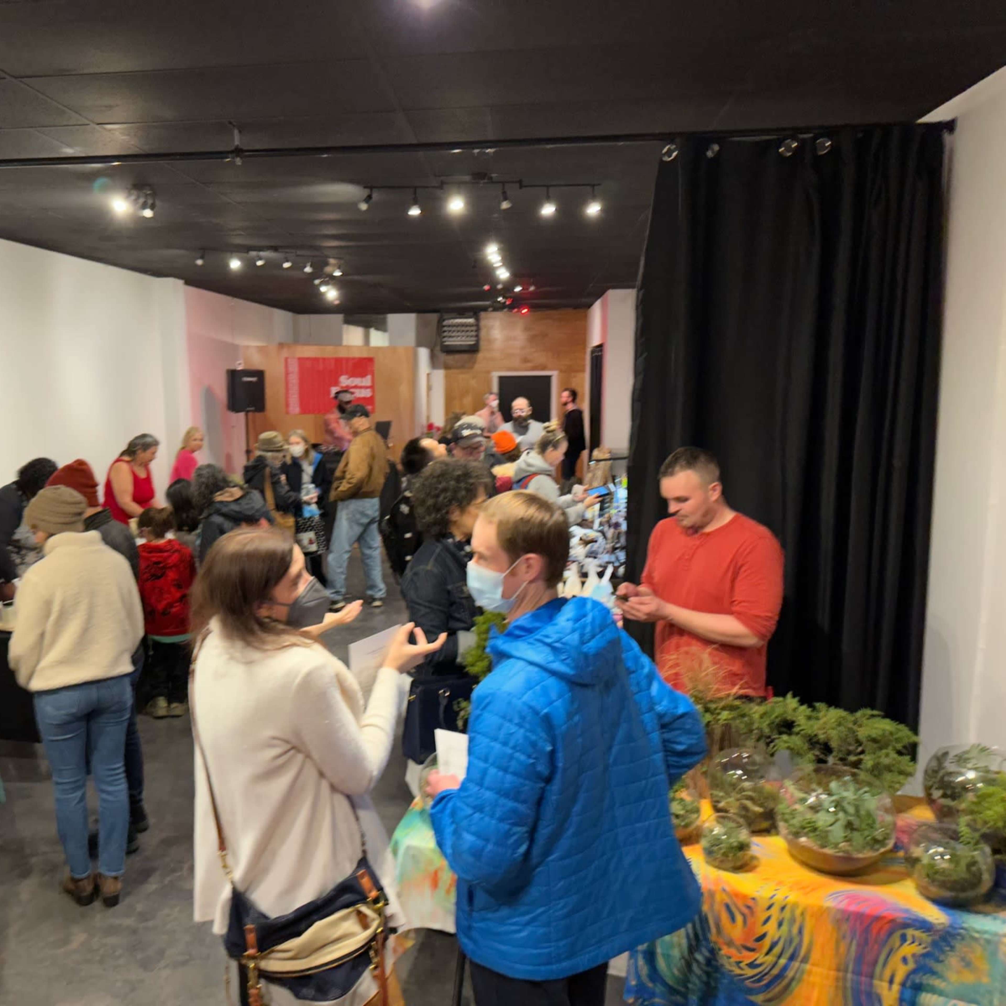 A group of people gathers in a spacious indoor area filled with tables, plants, and decorations, with some engaging in conversations and activities while a small stage is visible in the background.