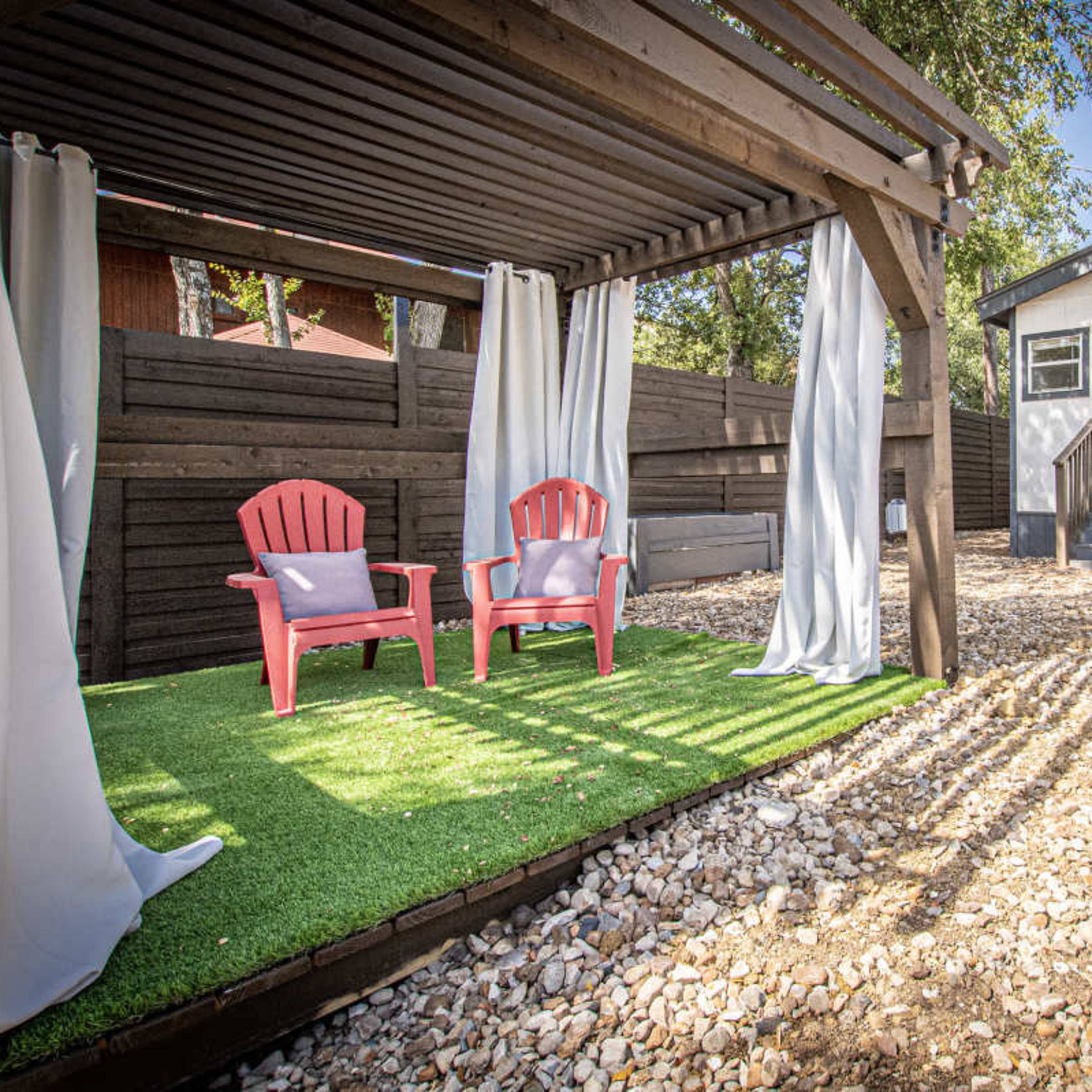 The scene features a wooden pergola with two red chairs and white curtains, surrounded by gravel and a small shed in a backyard setting.