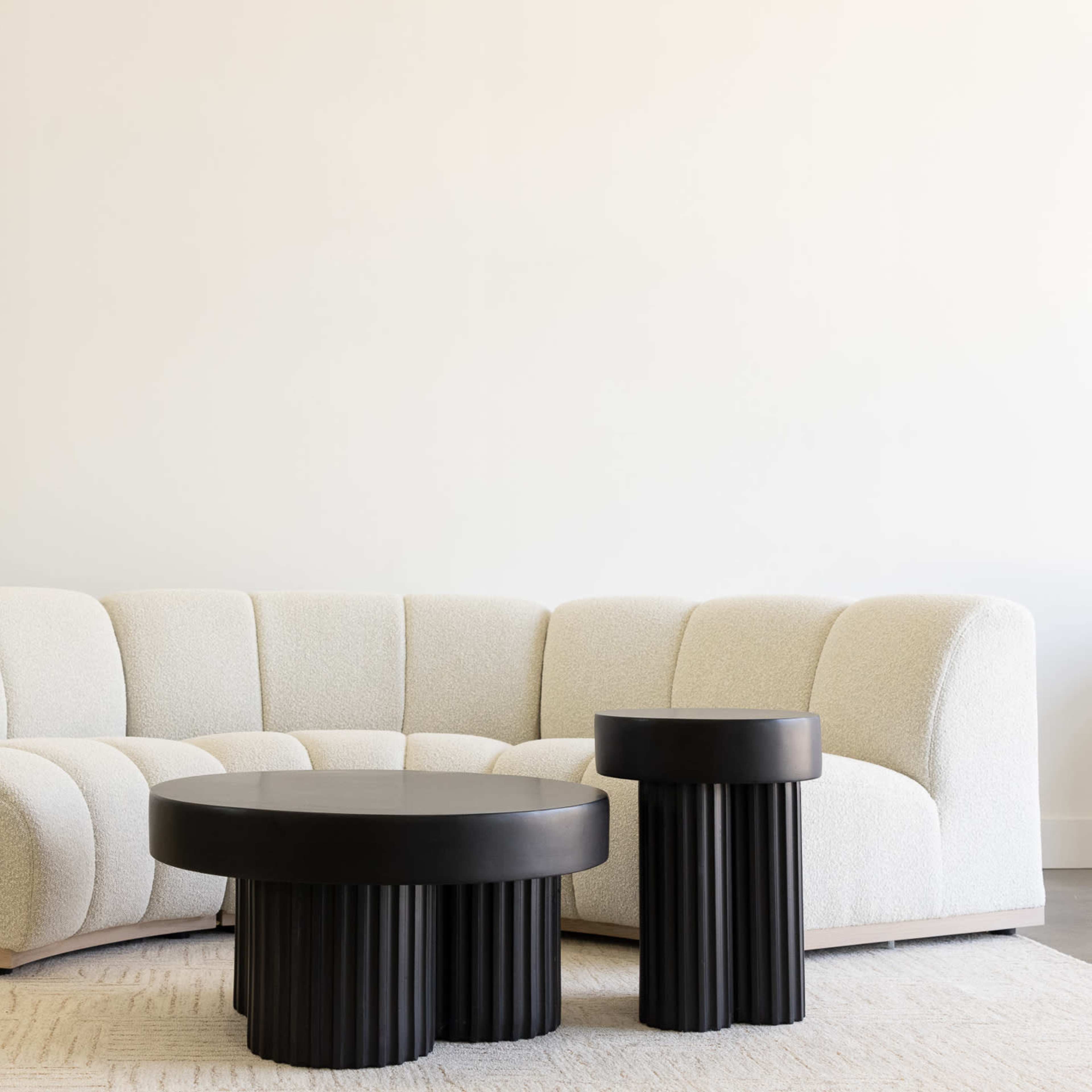 A curved, cream-colored sofa paired with two black, ribbed coffee tables on a light-colored rug in a minimalist interior.