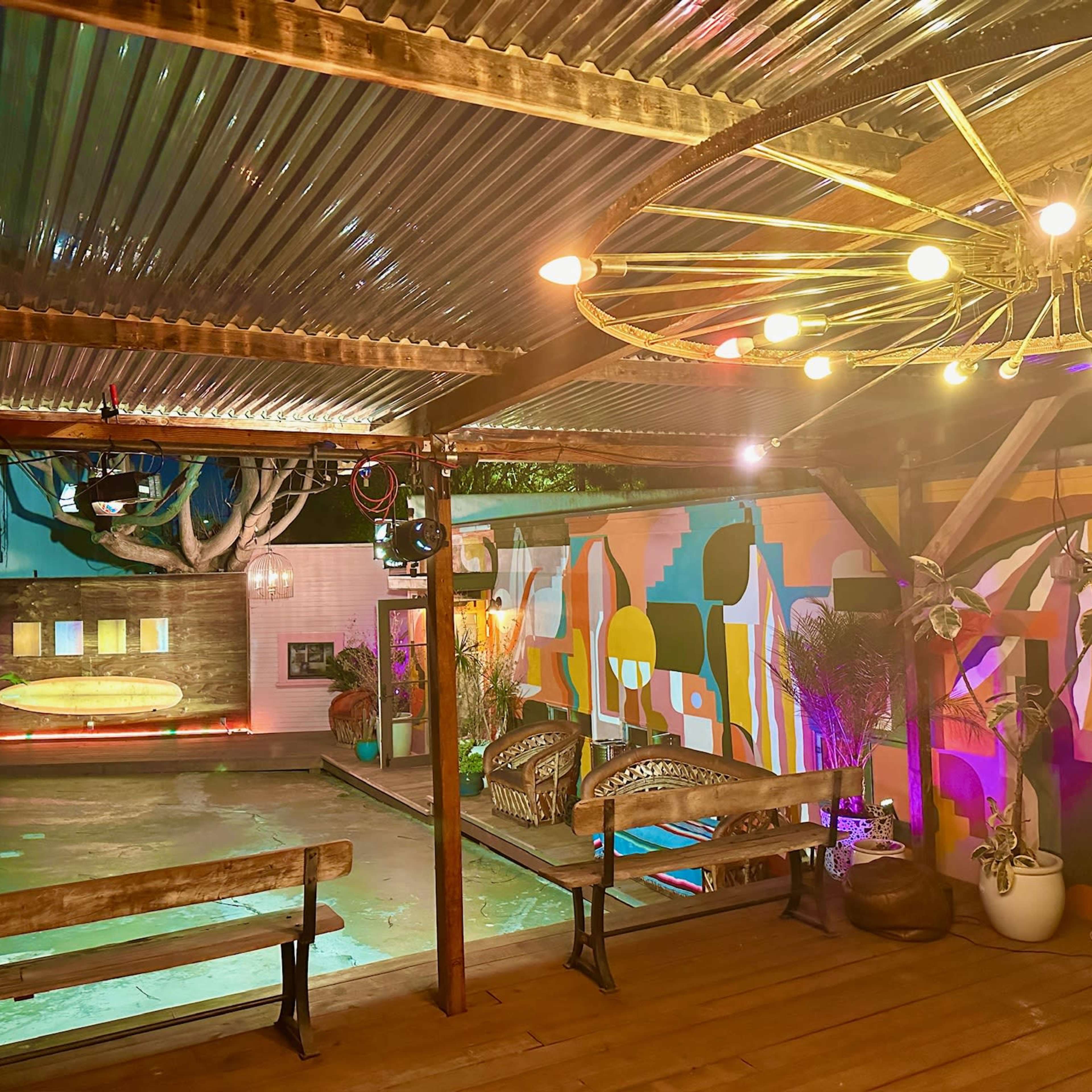 A colorful outdoor space with a pool, wooden benches, and decorative lights hanging from a metal roof.