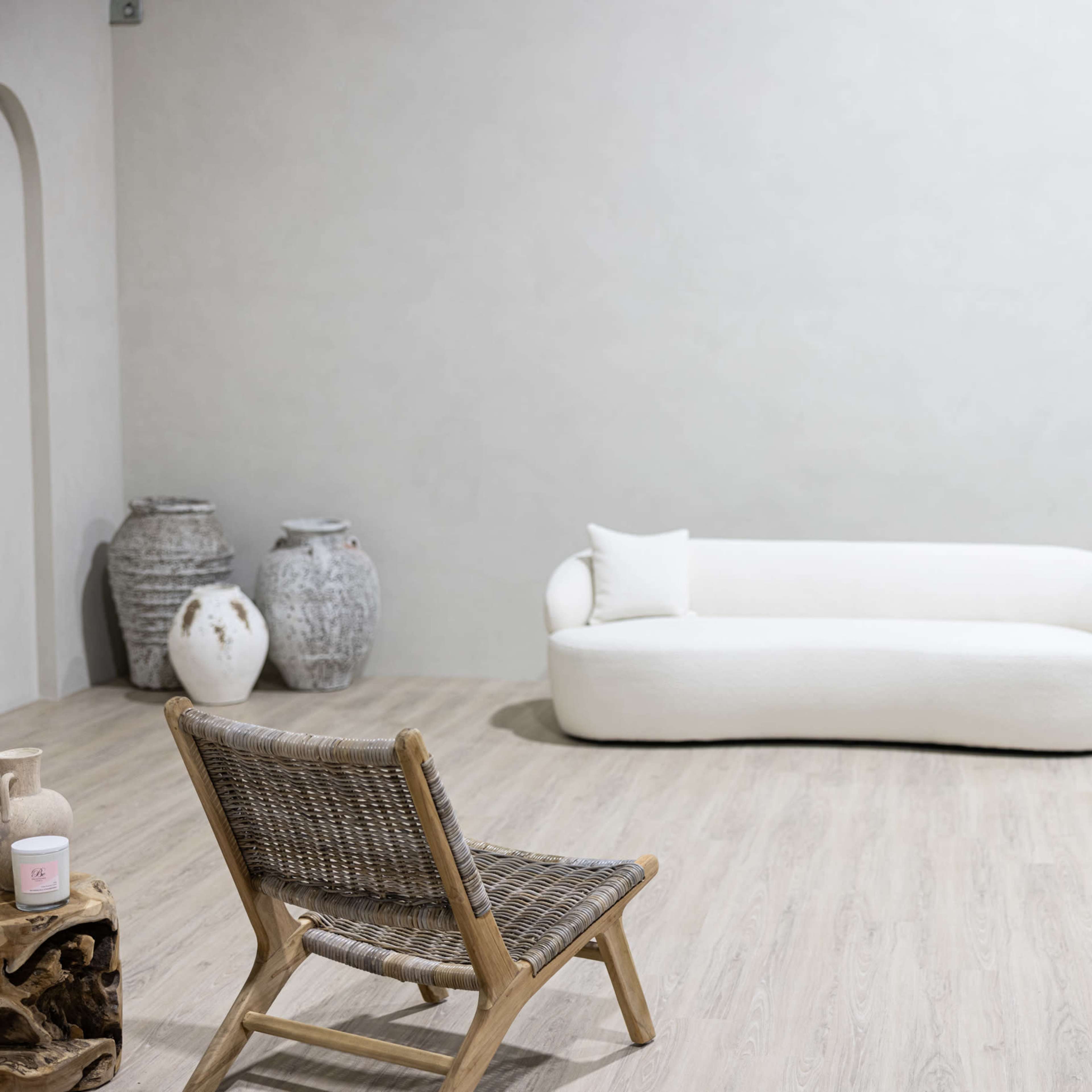 A minimalist interior space with a white sofa, a wooden chair, decorative vases, and an arched mirror against a light-colored wall.