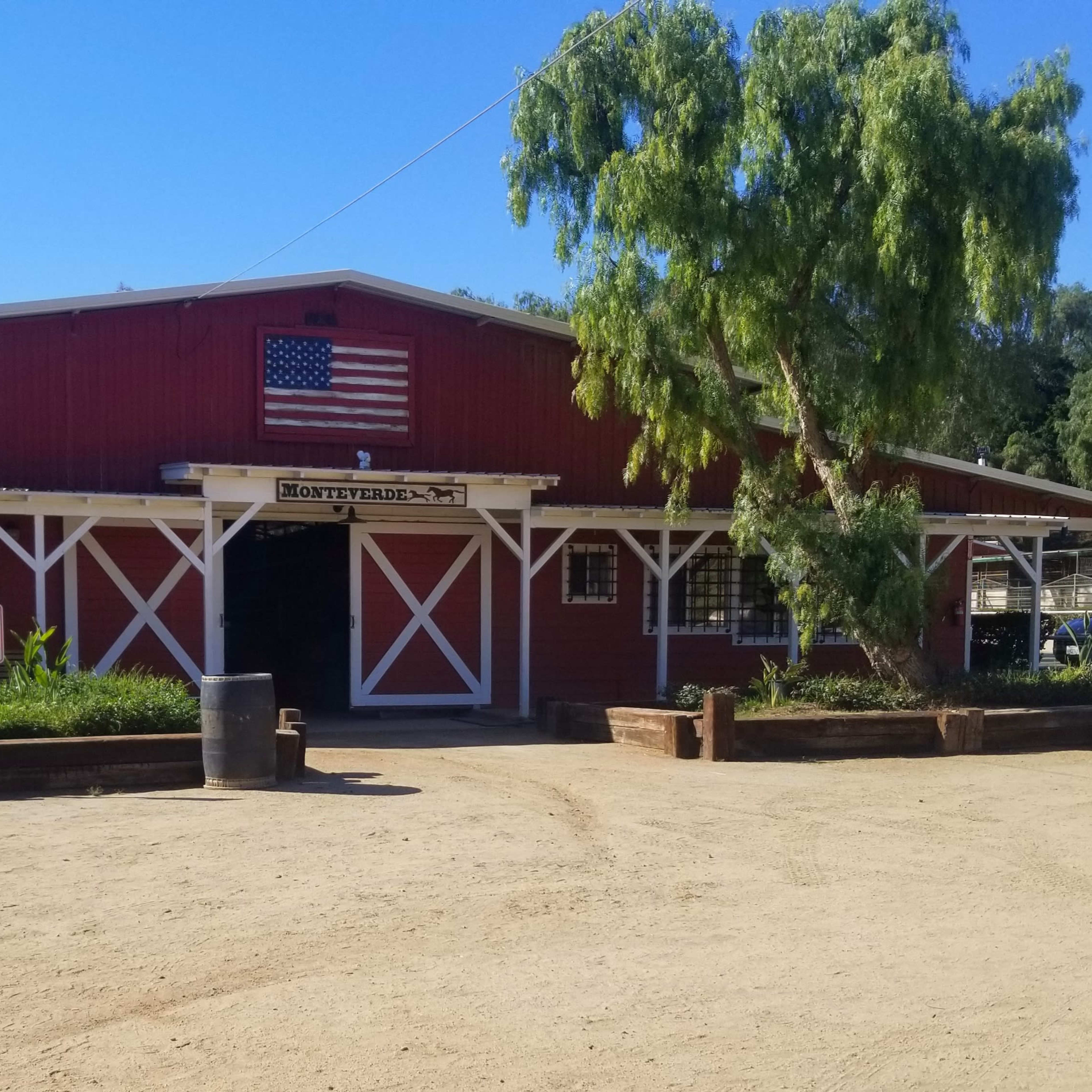A red barn with a flag on the front is surrounded by greenery and features a gravel path leading to the entrance.