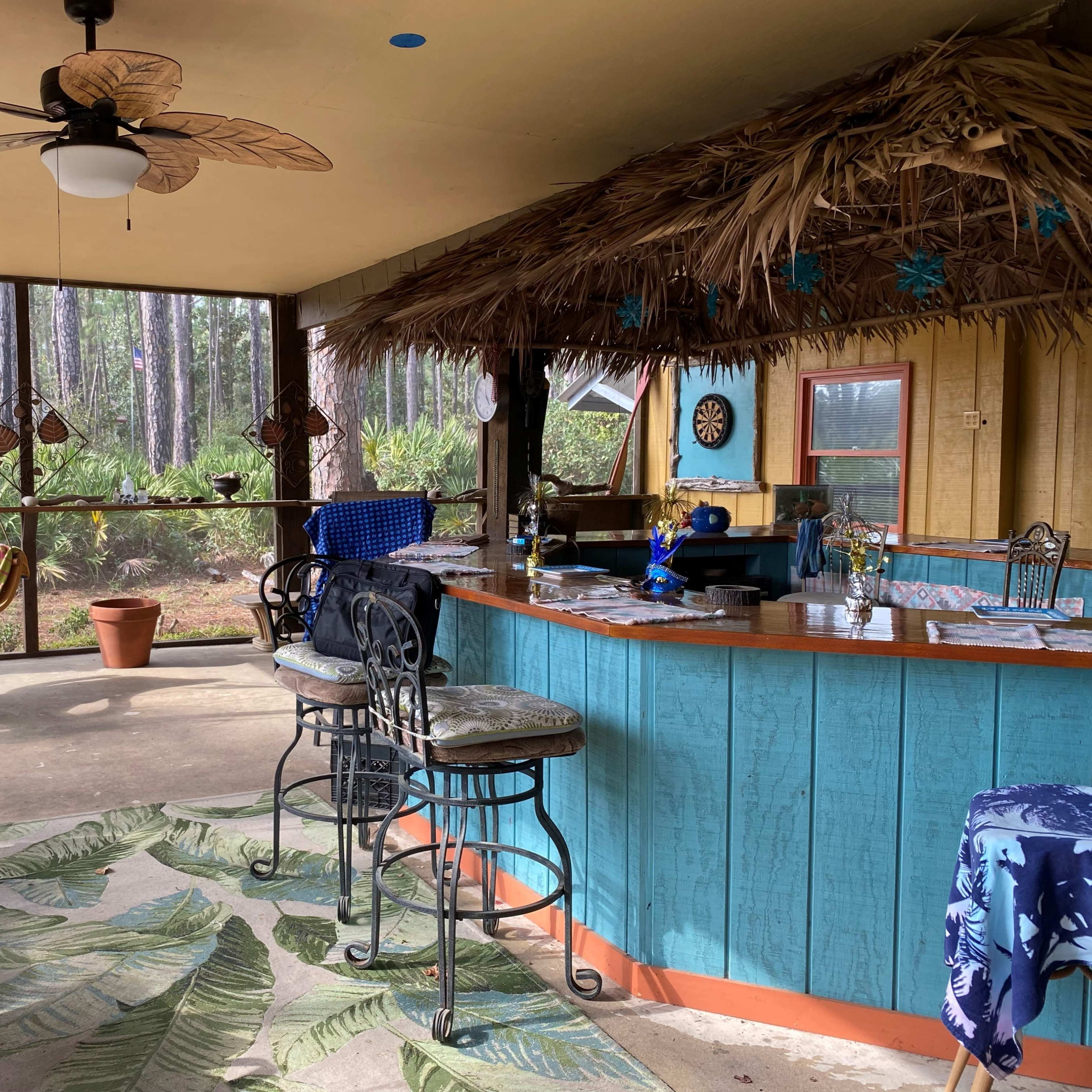 A screened-in porch featuring a bar with high stools, tropical-themed decor, and seating arrangements under a thatched roof.