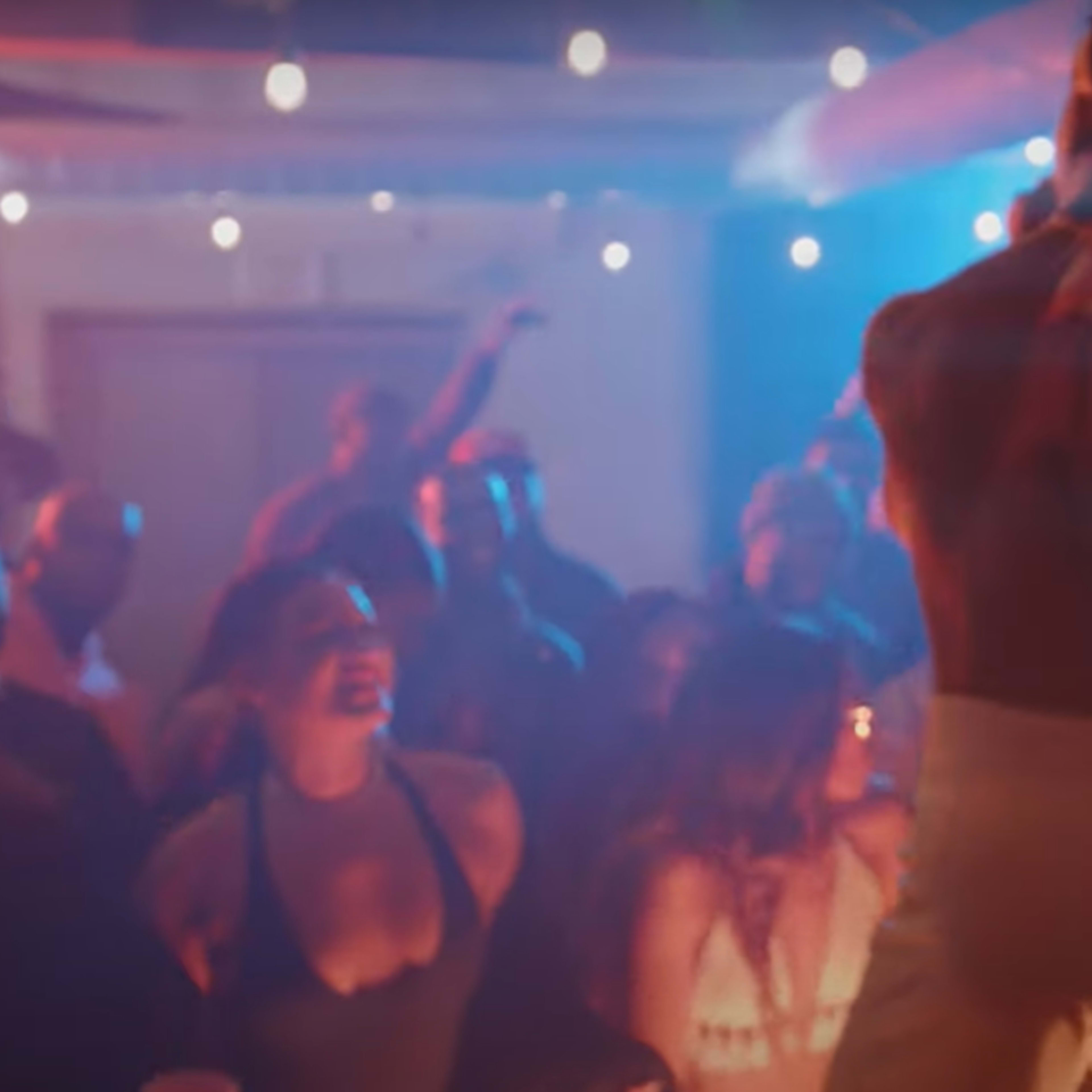 A performer stands on stage with their back to the audience, surrounded by a lively crowd dancing and enjoying the atmosphere of a dimly lit venue.