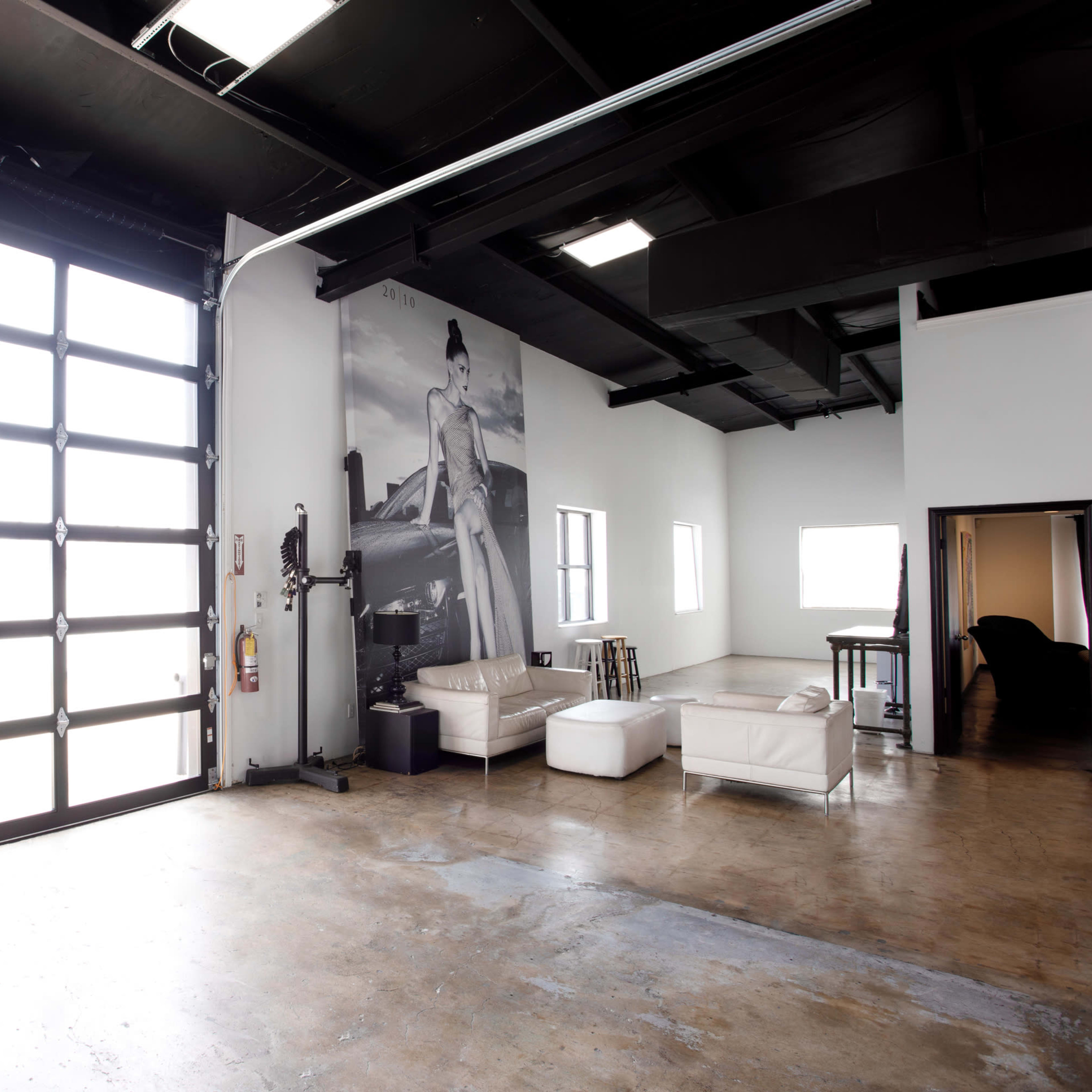 A spacious room with large windows, a rolled-up garage door, and minimal furniture including white seating and a table.