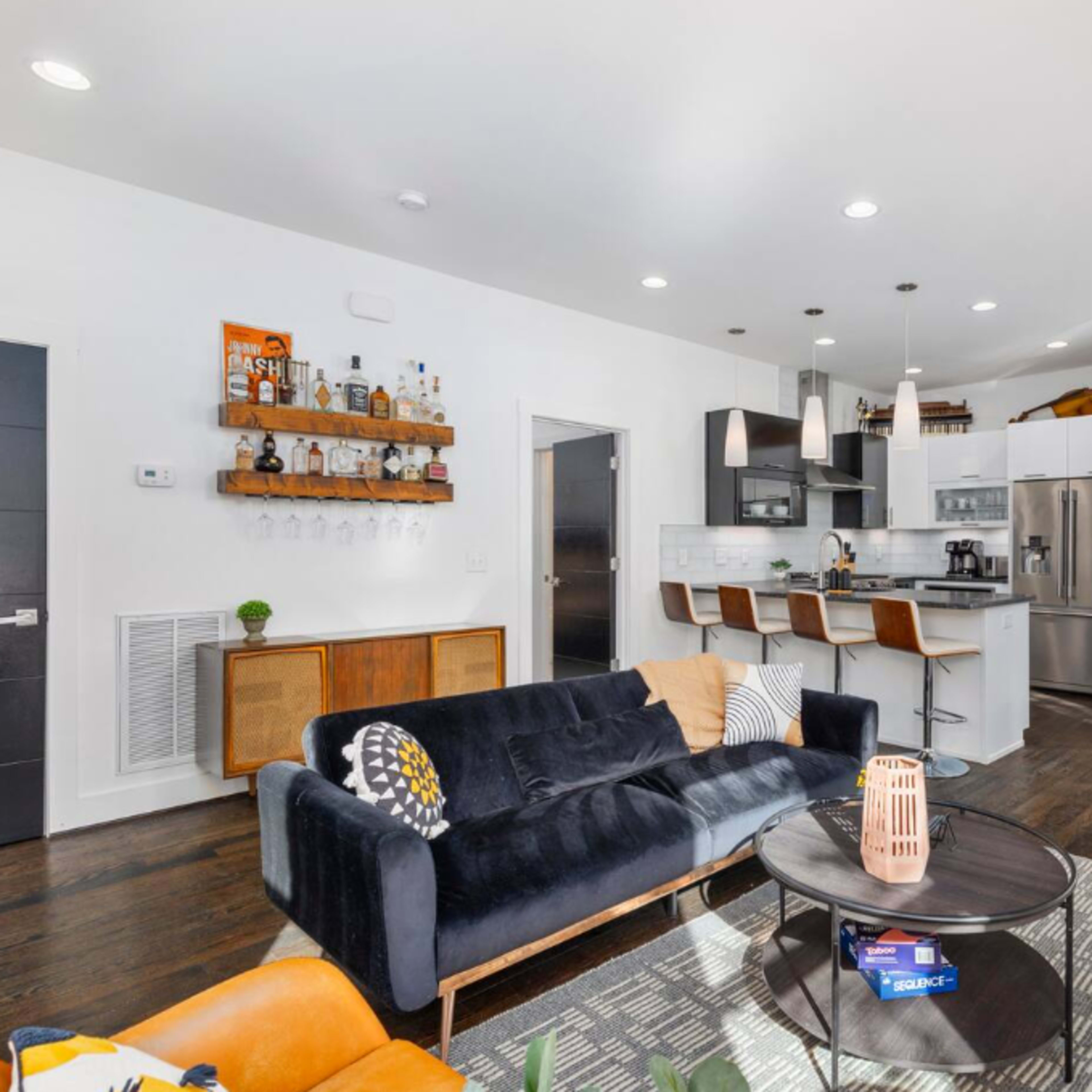 A modern living room with a black sofa, a round coffee table, bar stools at a kitchen counter, and a staircase leading to the upper level.