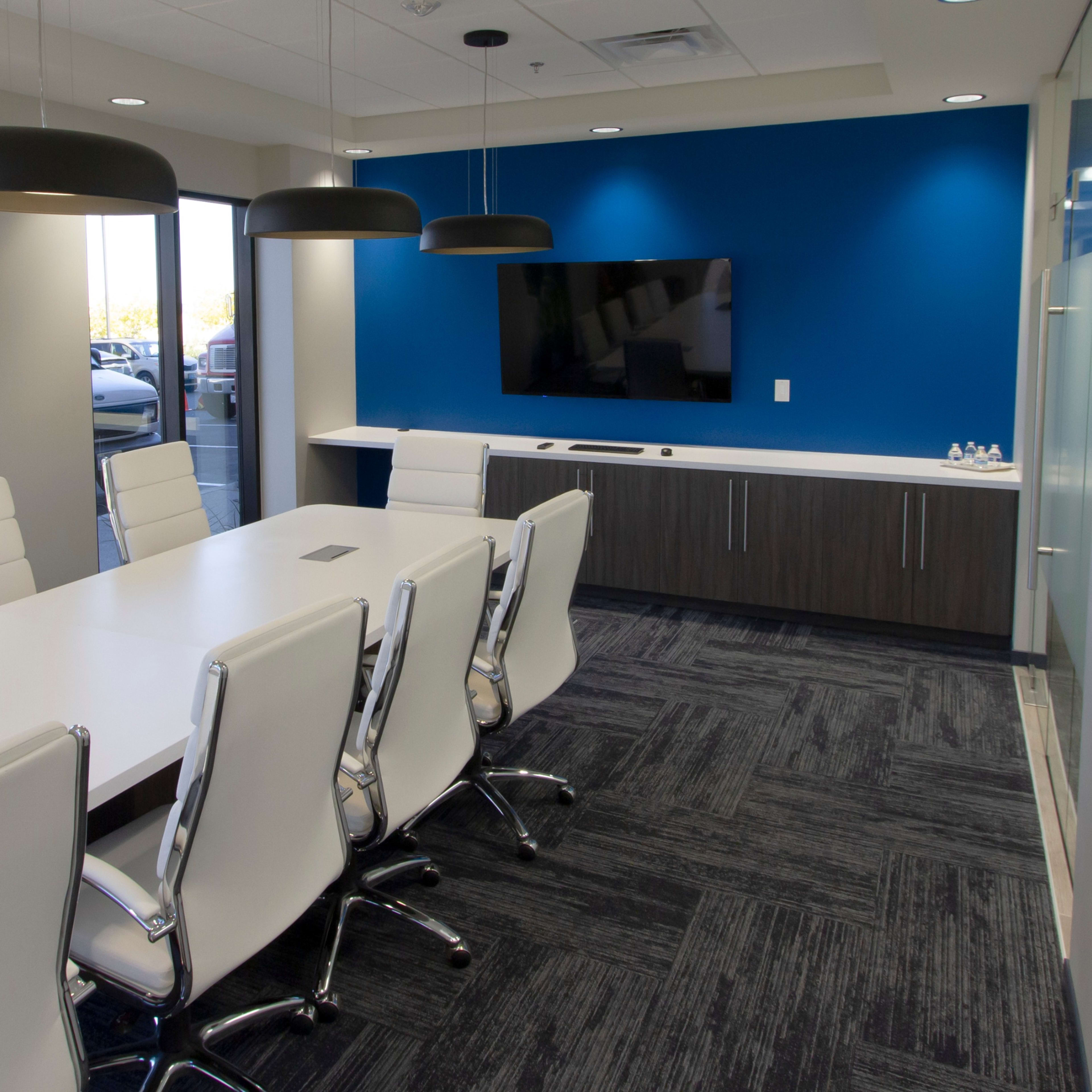 A modern conference room features a long white table surrounded by sleek white chairs, with a television mounted on a blue accent wall.
