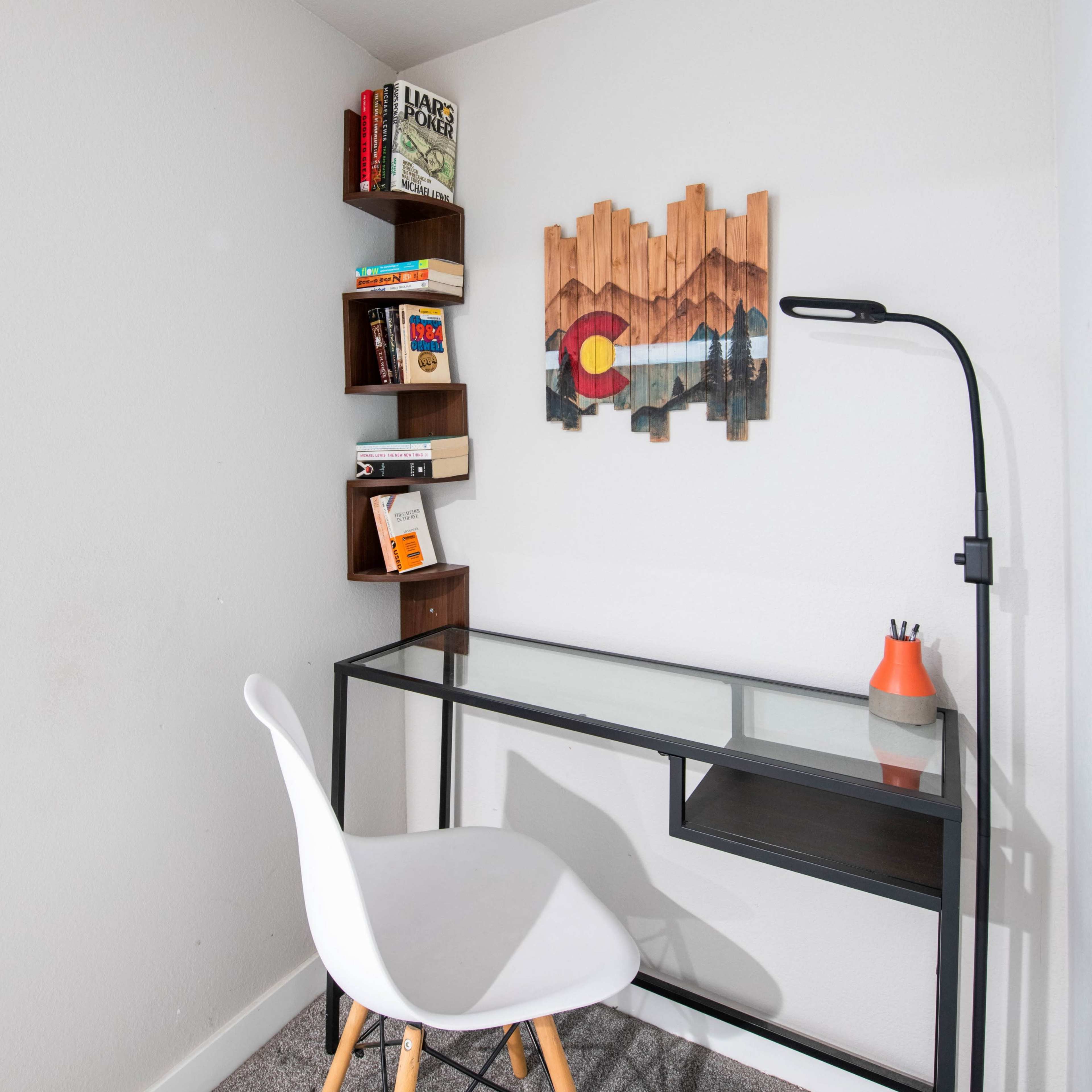 A small workspace features a glass-top desk, a white chair, a bookshelf with several books, and a wall art piece depicting a mountain landscape with a Colorado flag motif.