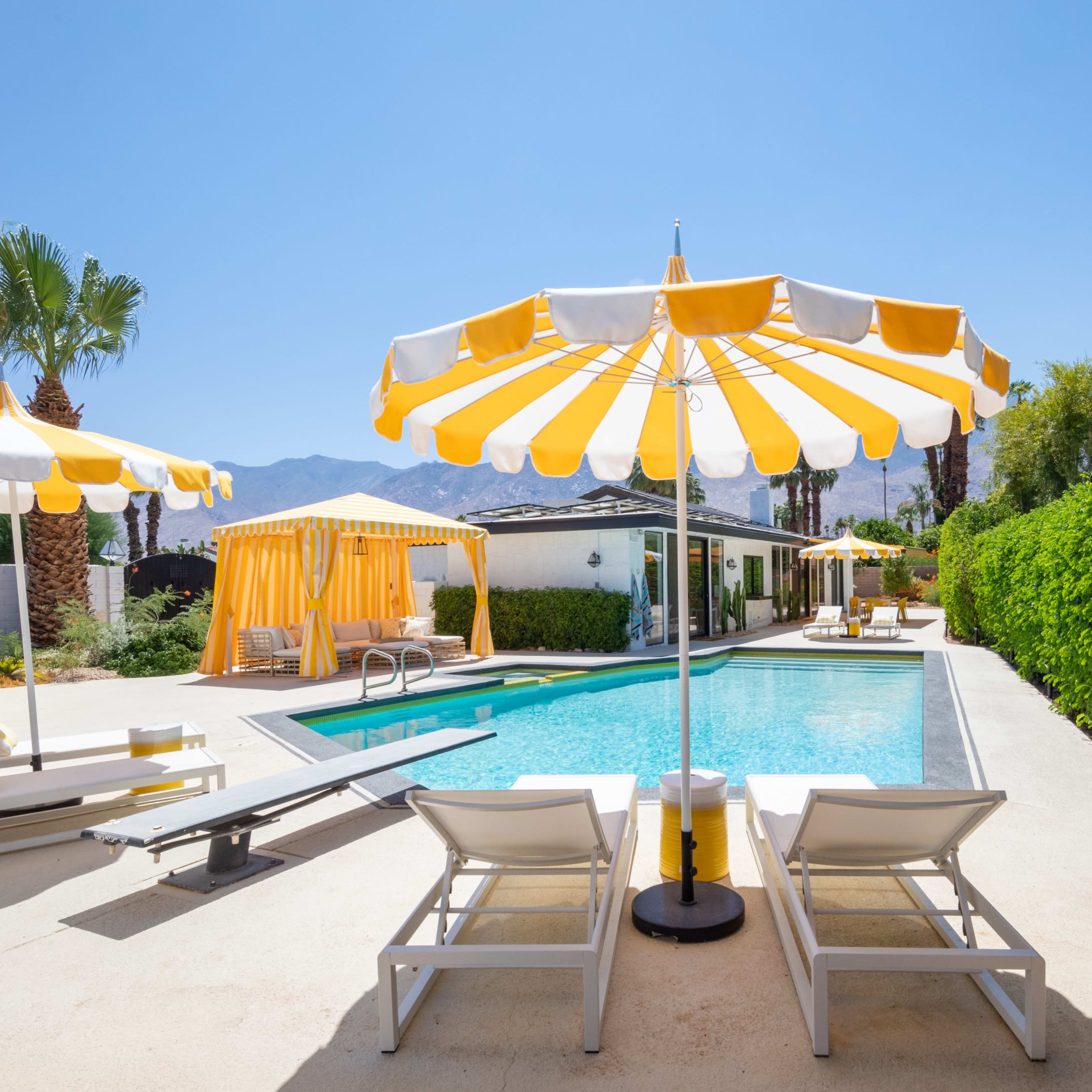 A pool area features lounge chairs and yellow and white umbrellas, surrounded by green hedges and palm trees under a clear blue sky.