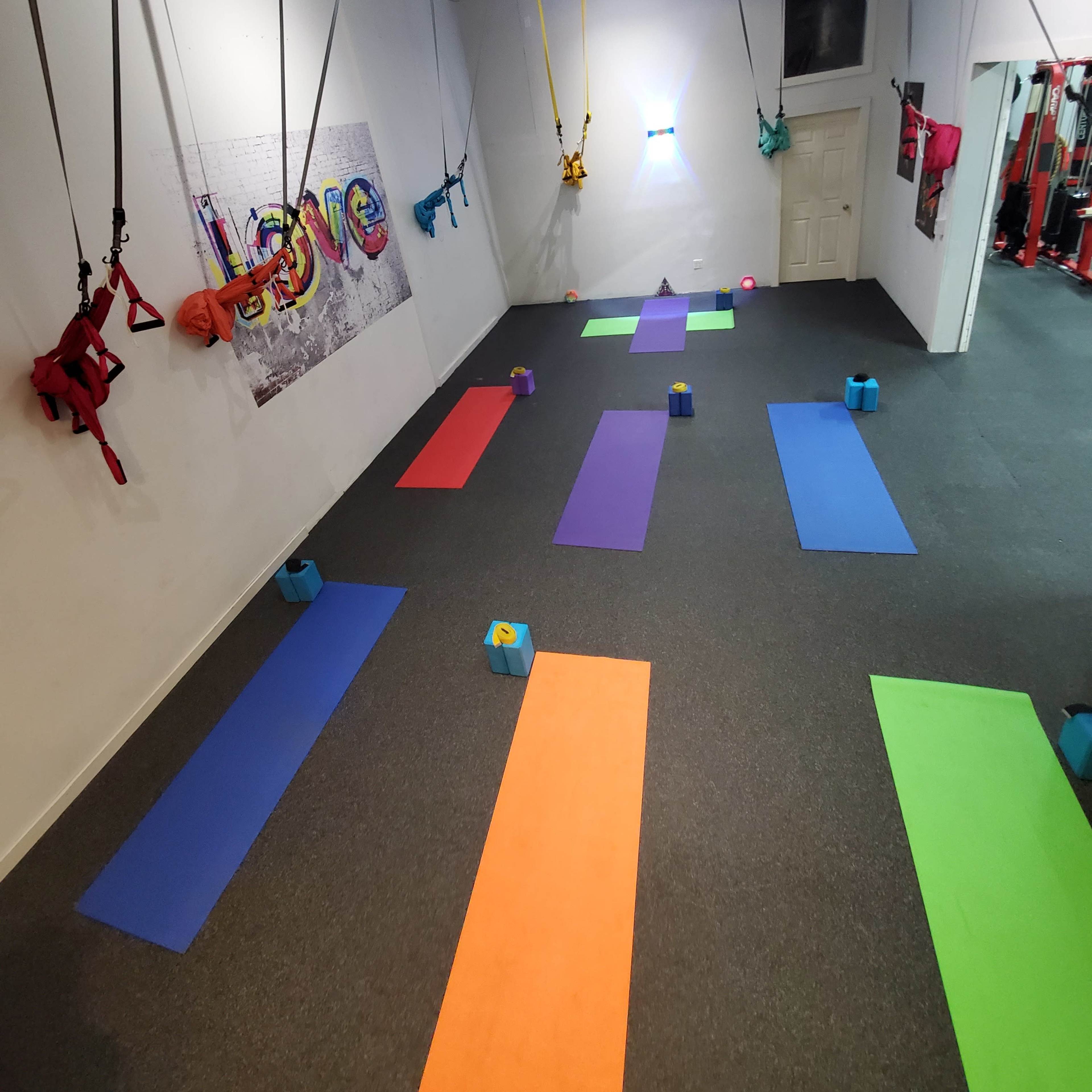 A brightly colored fitness studio with various exercise mats arranged on the floor and harnesses suspended from the ceiling.