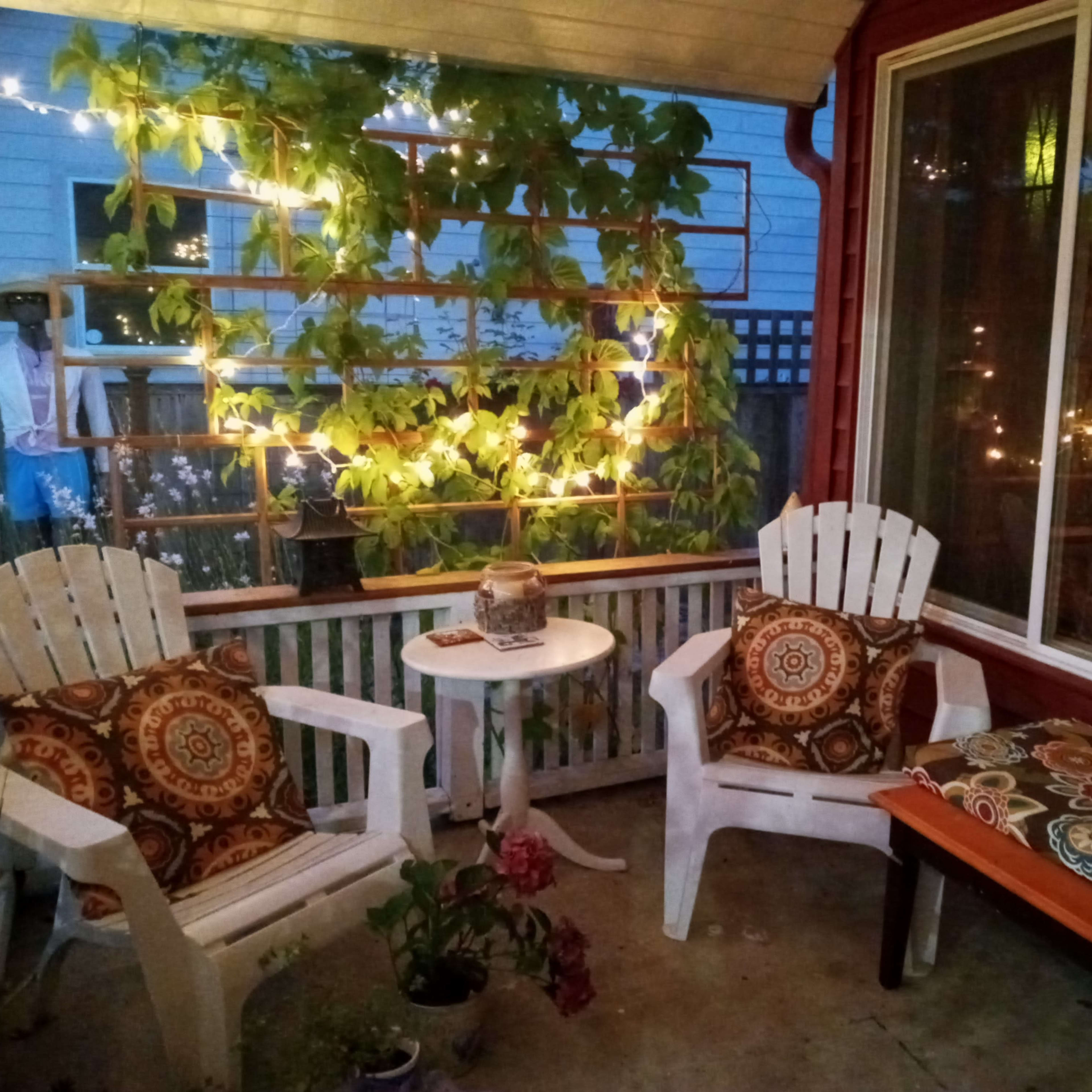 A cozy porch features two white adirondack chairs with patterned cushions, a small table, and greenery illuminated by string lights.