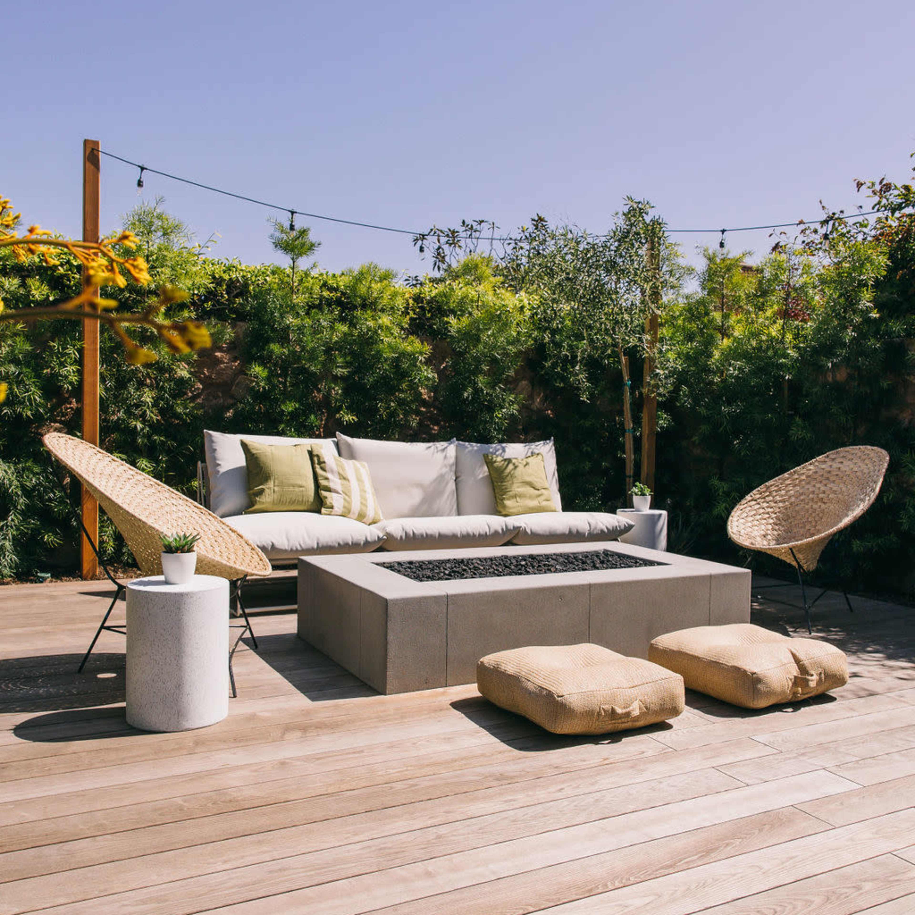 A stylish outdoor seating area with a fire pit, surrounded by greenery and featuring comfortable lounge chairs and cushions.