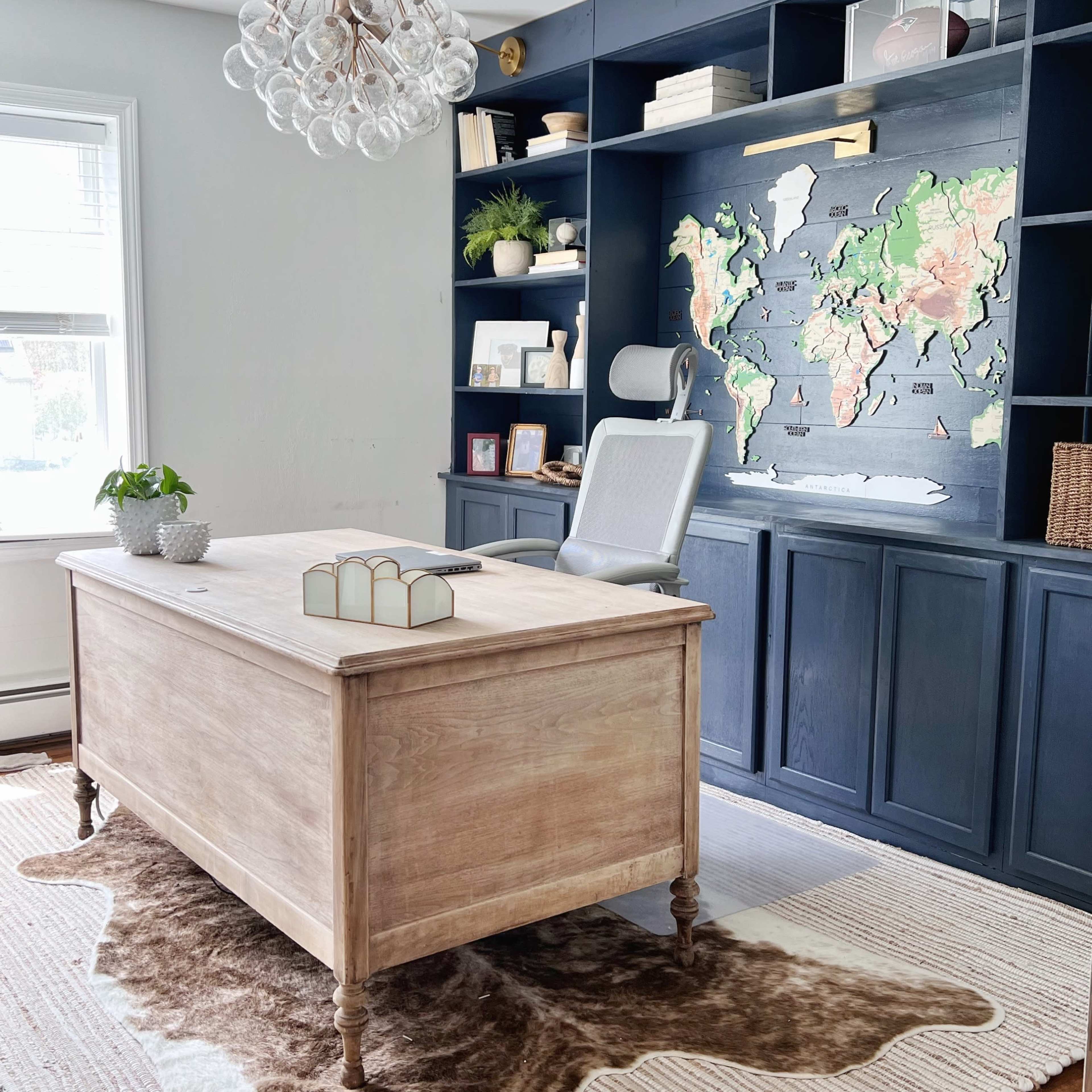 A home office with a wooden desk, a white ergonomic chair, a map on the wall, and built-in shelves displaying decor items.
