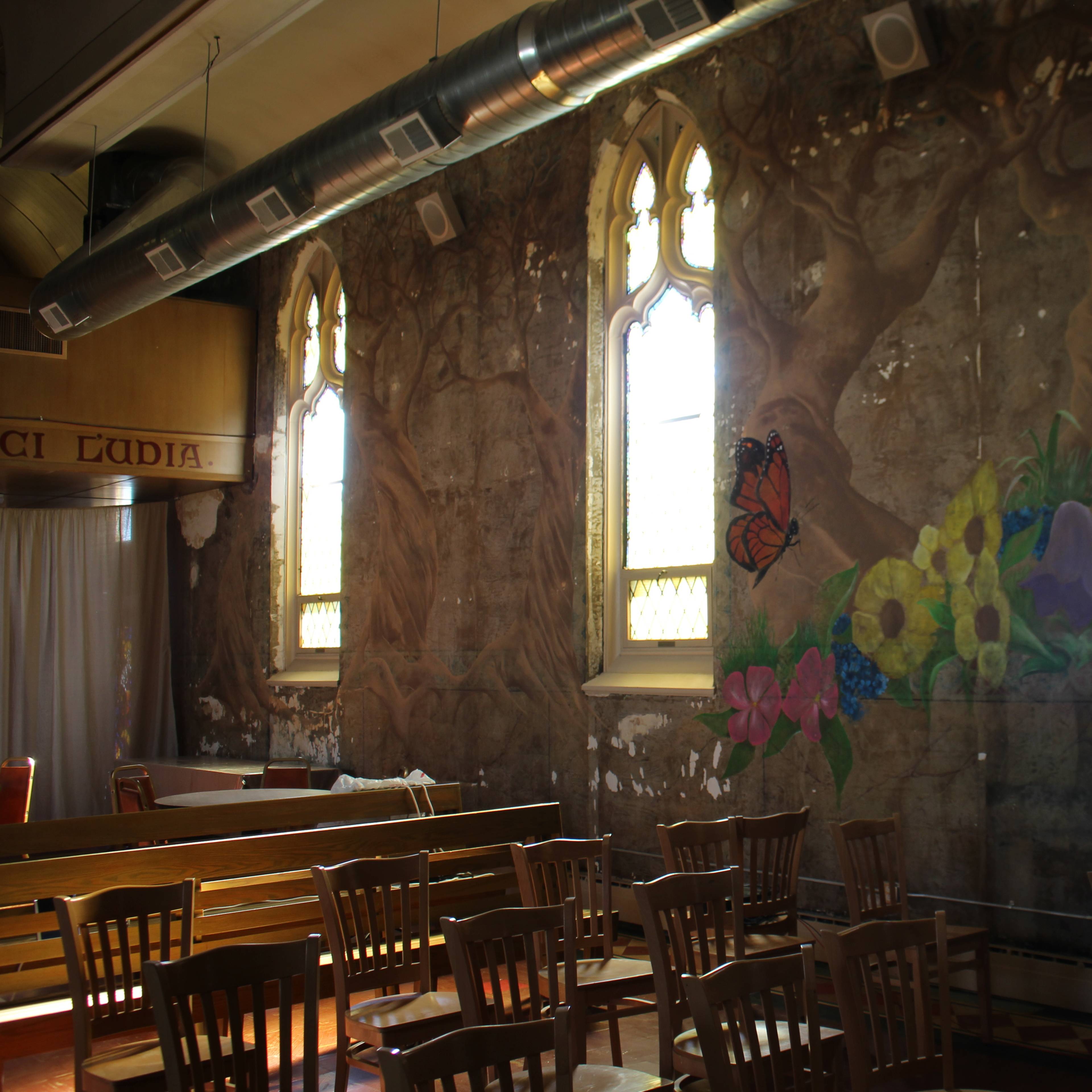An interior space with wooden chairs arranged around a wall featuring faded murals and large, arched windows.