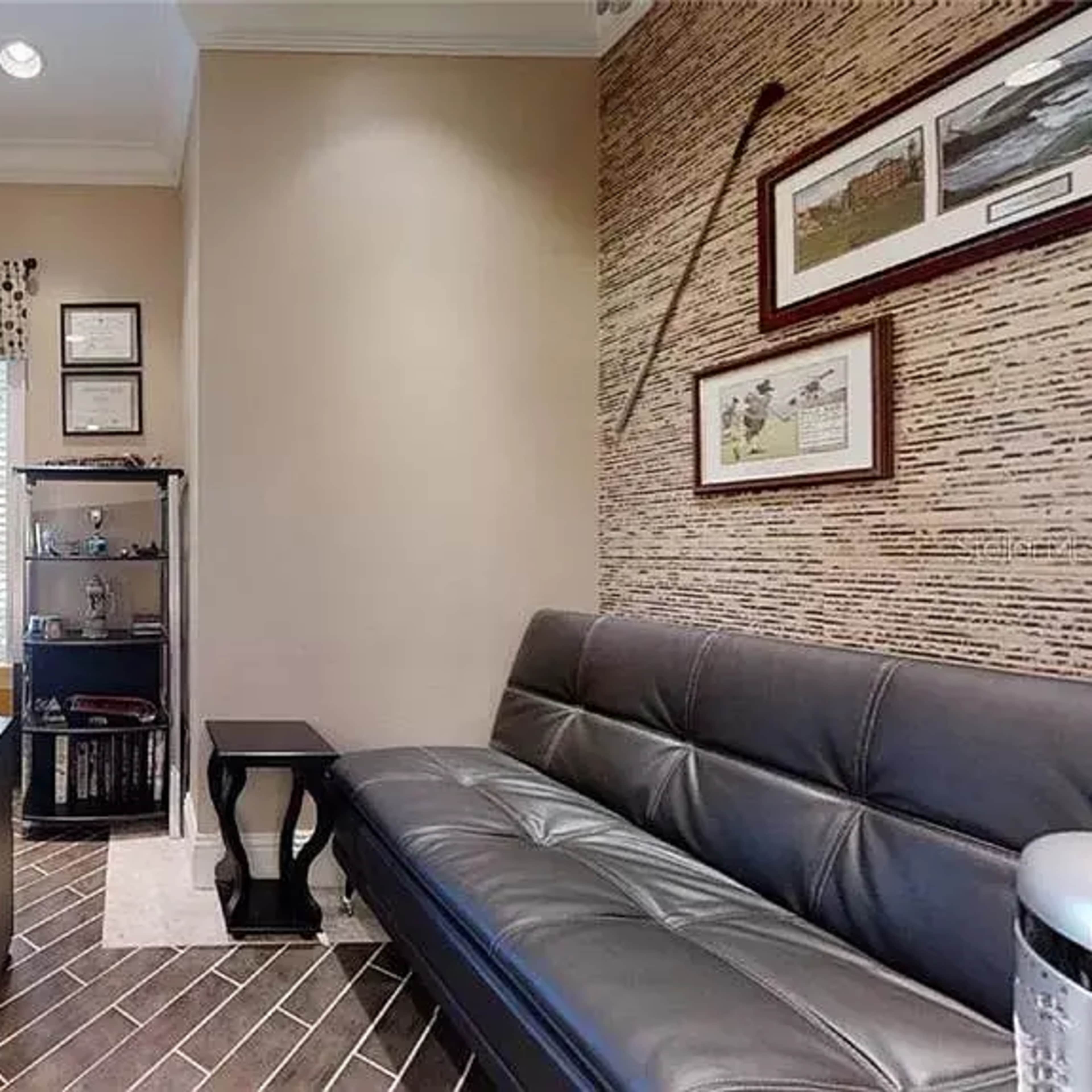 A small office with a desk, chair, and a black couch, featuring a window with a patterned curtain and a wall decorated with framed pictures.