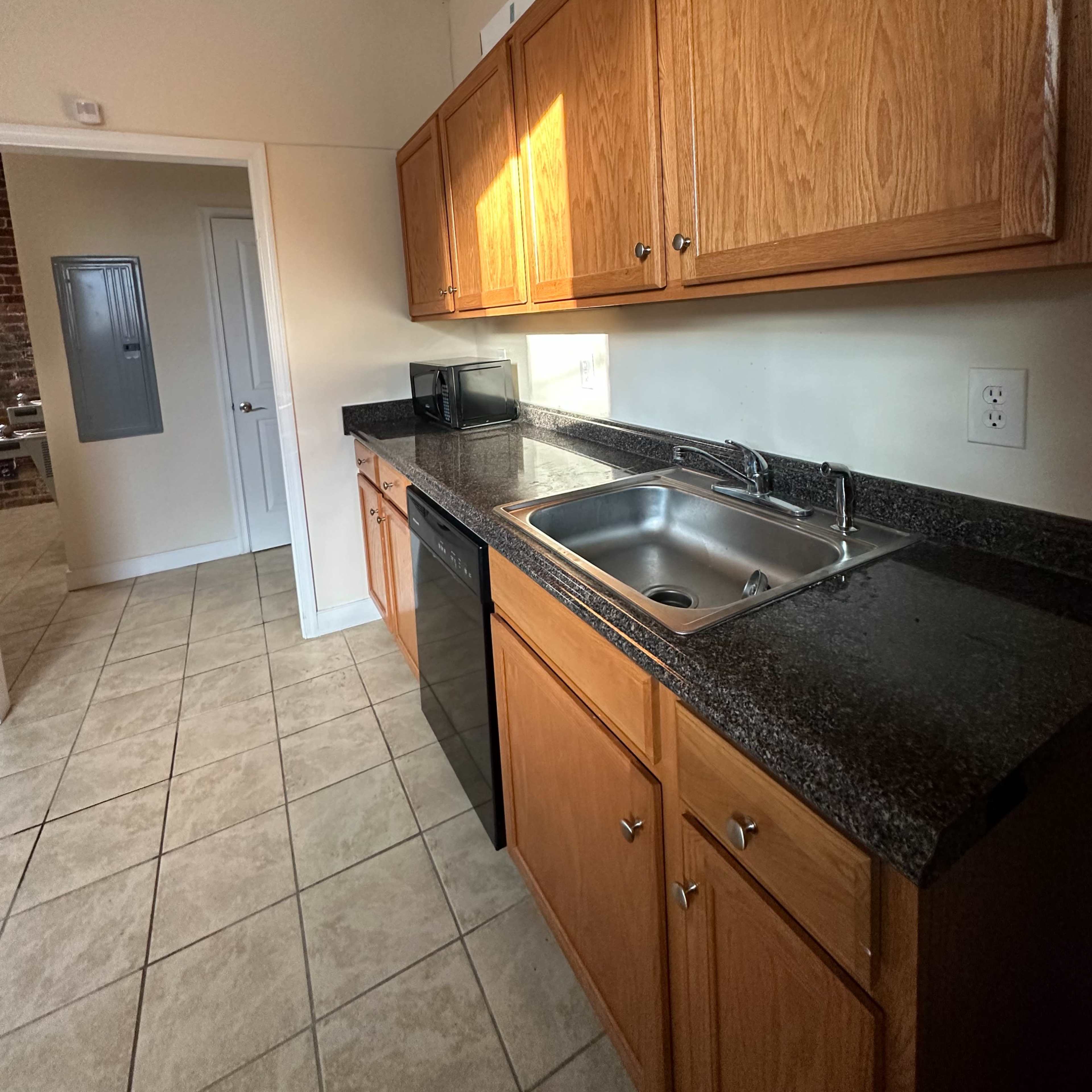 A kitchen with wooden cabinetry, a stainless steel sink, and dark countertops, featuring tile flooring and a doorway leading to another room.