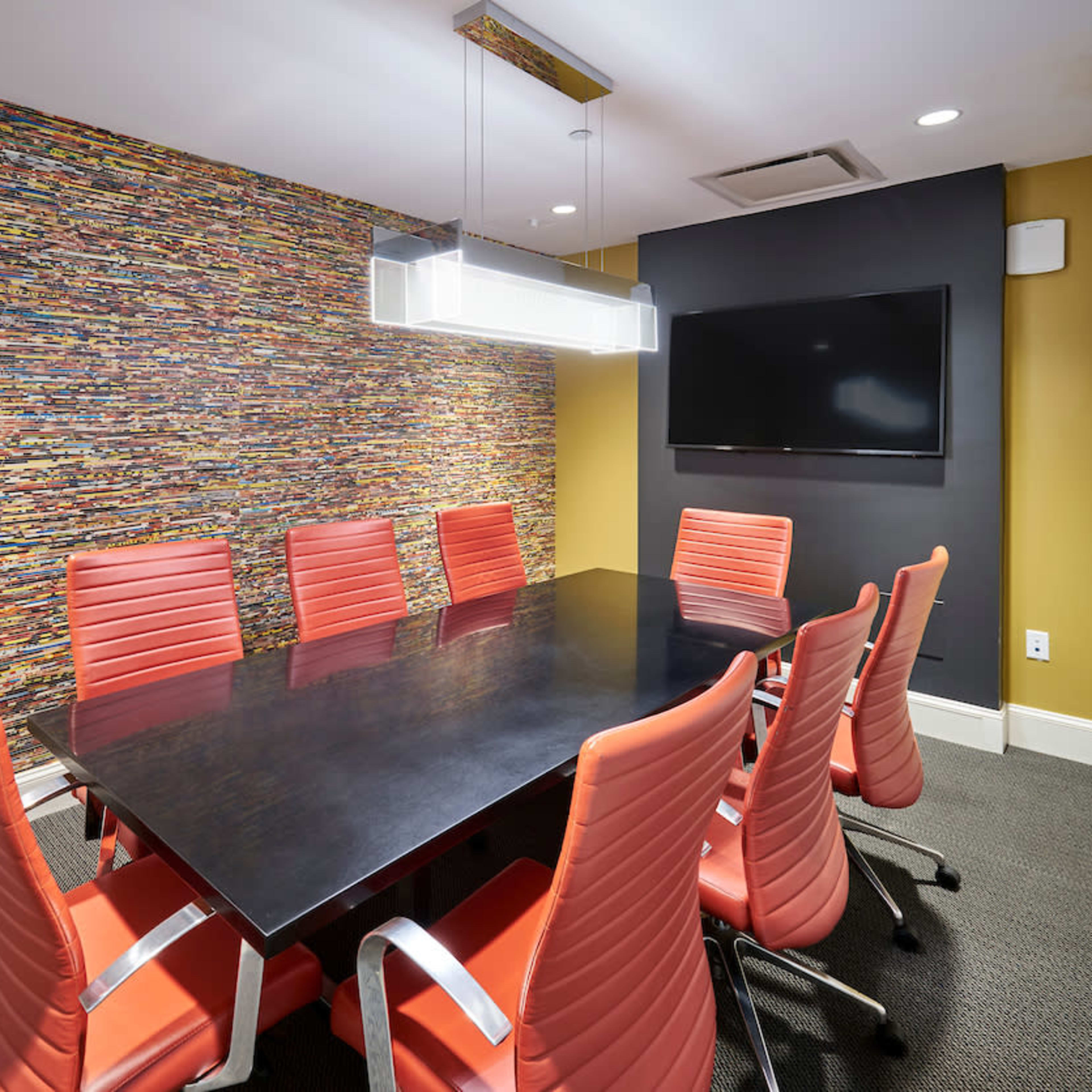 A modern conference room features a long black table surrounded by orange chairs, with a colorful patterned wall and a flat-screen TV.