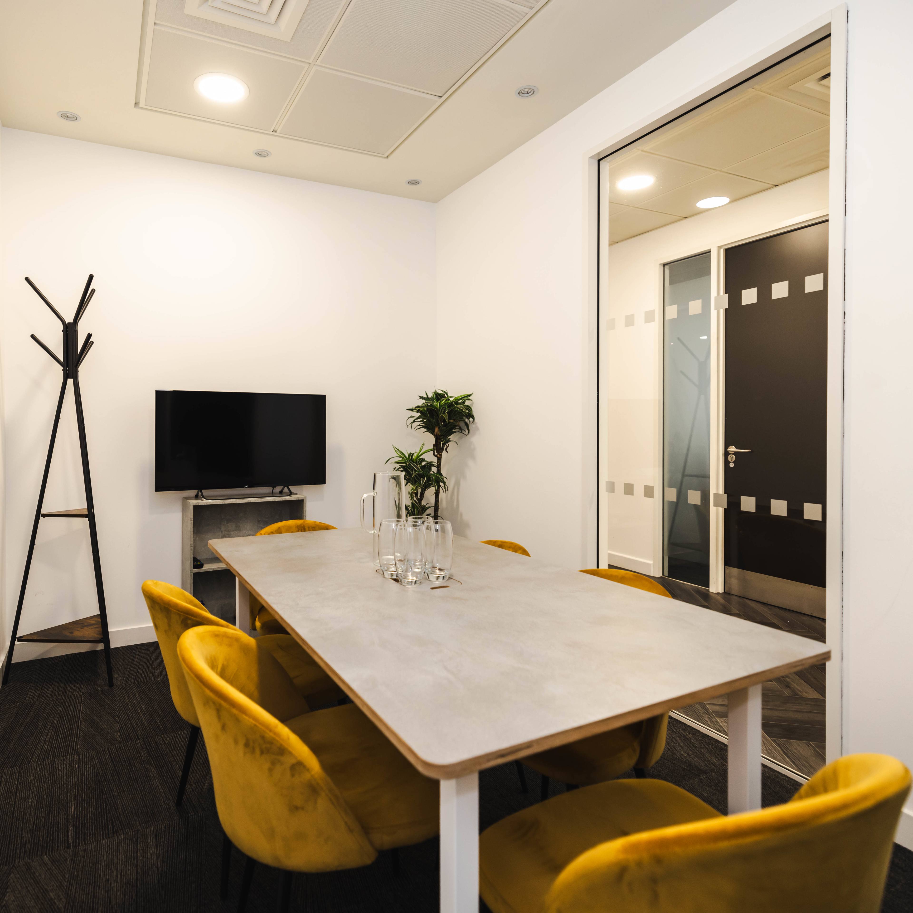 A small meeting room features a gray table surrounded by yellow chairs, a television mounted on the wall, and a plant beside a coat rack.