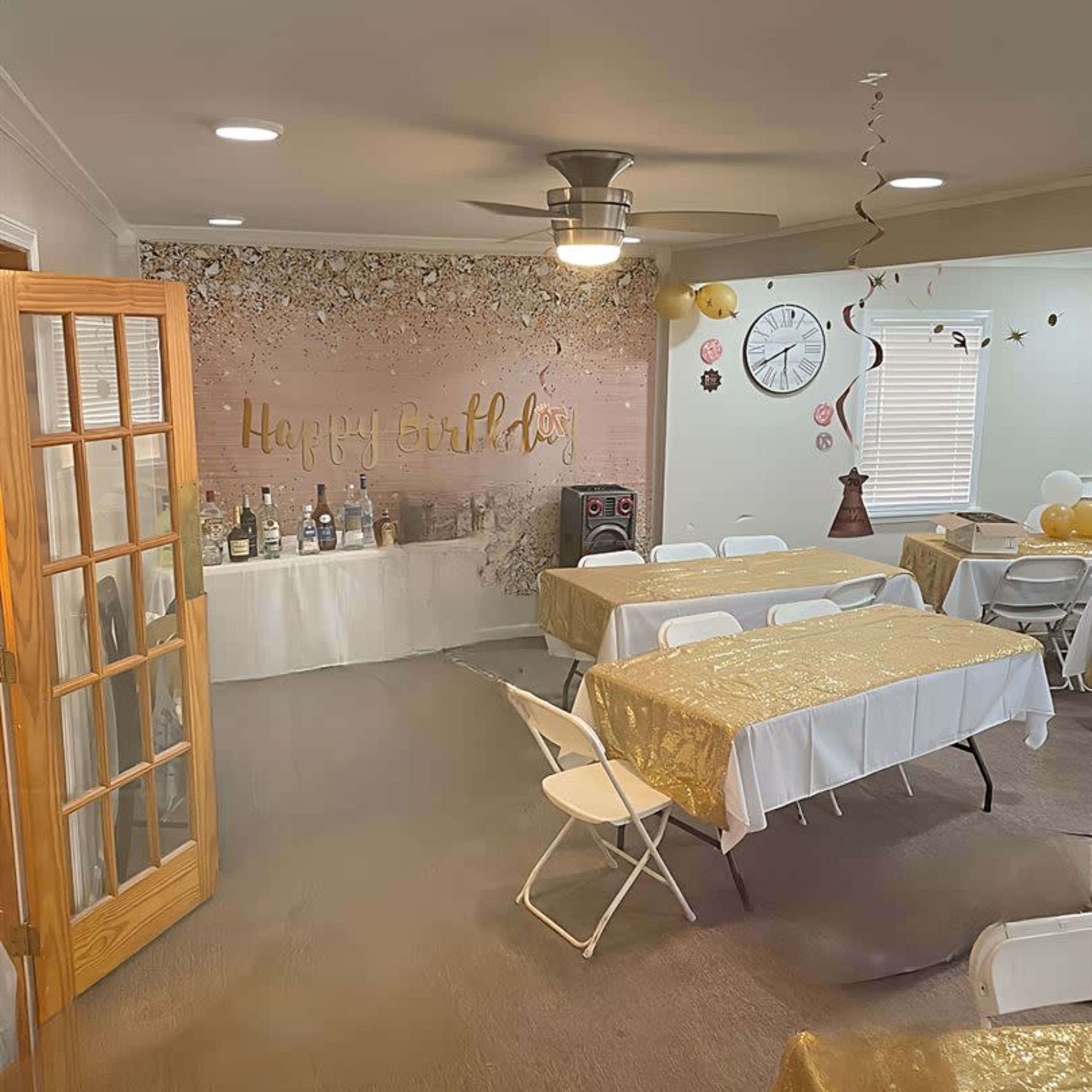 A decorated indoor space for a birthday celebration, featuring tables with gold tablecloths, a "Happy Birthday" sign on the wall, and various bottles arranged on a display table.