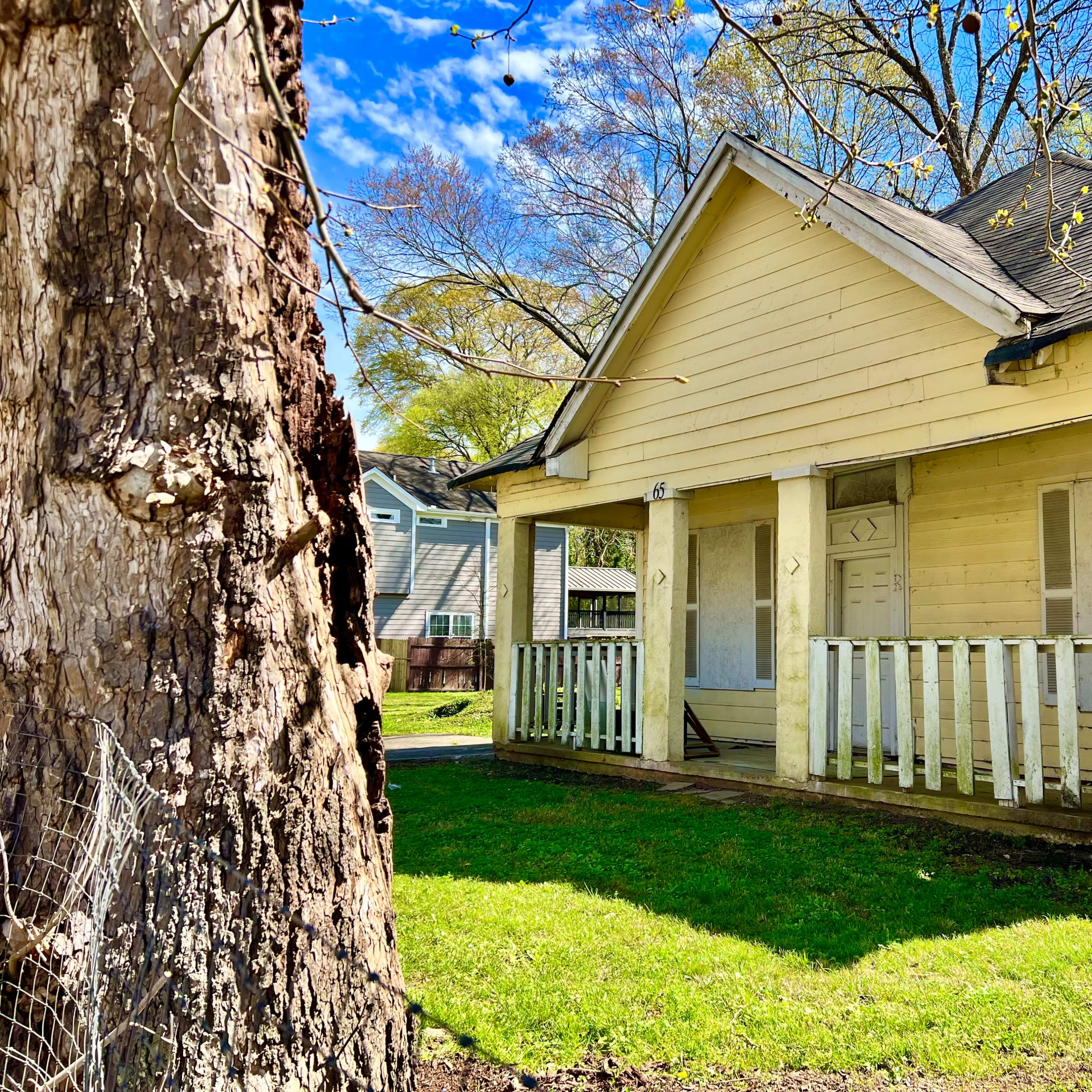 A yellow wooden house with a porch is situated next to a large tree in a grassy area.