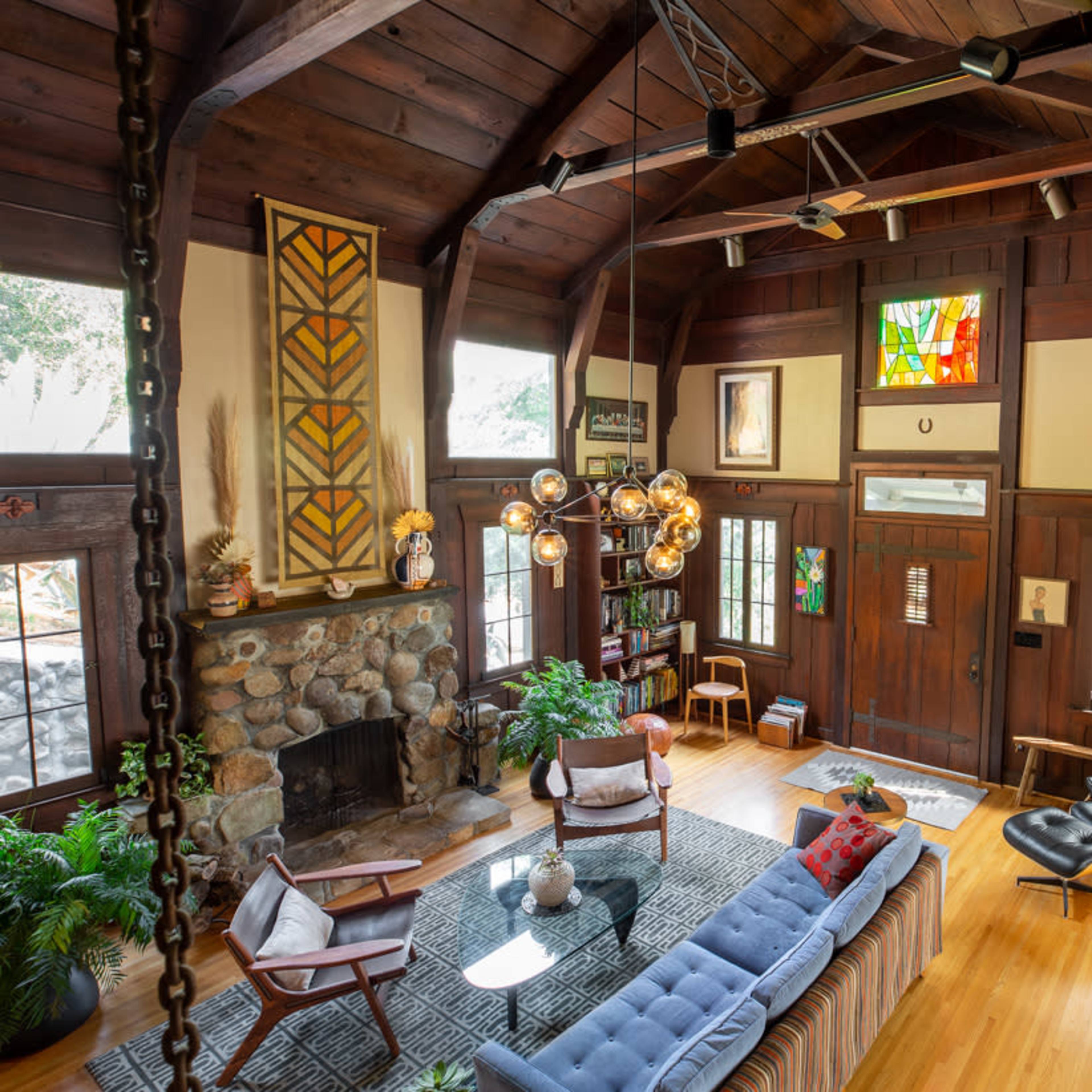 A spacious living room with a stone fireplace, wooden beams, and a mix of modern and vintage furniture, including a blue sofa and an art-decorated wall.