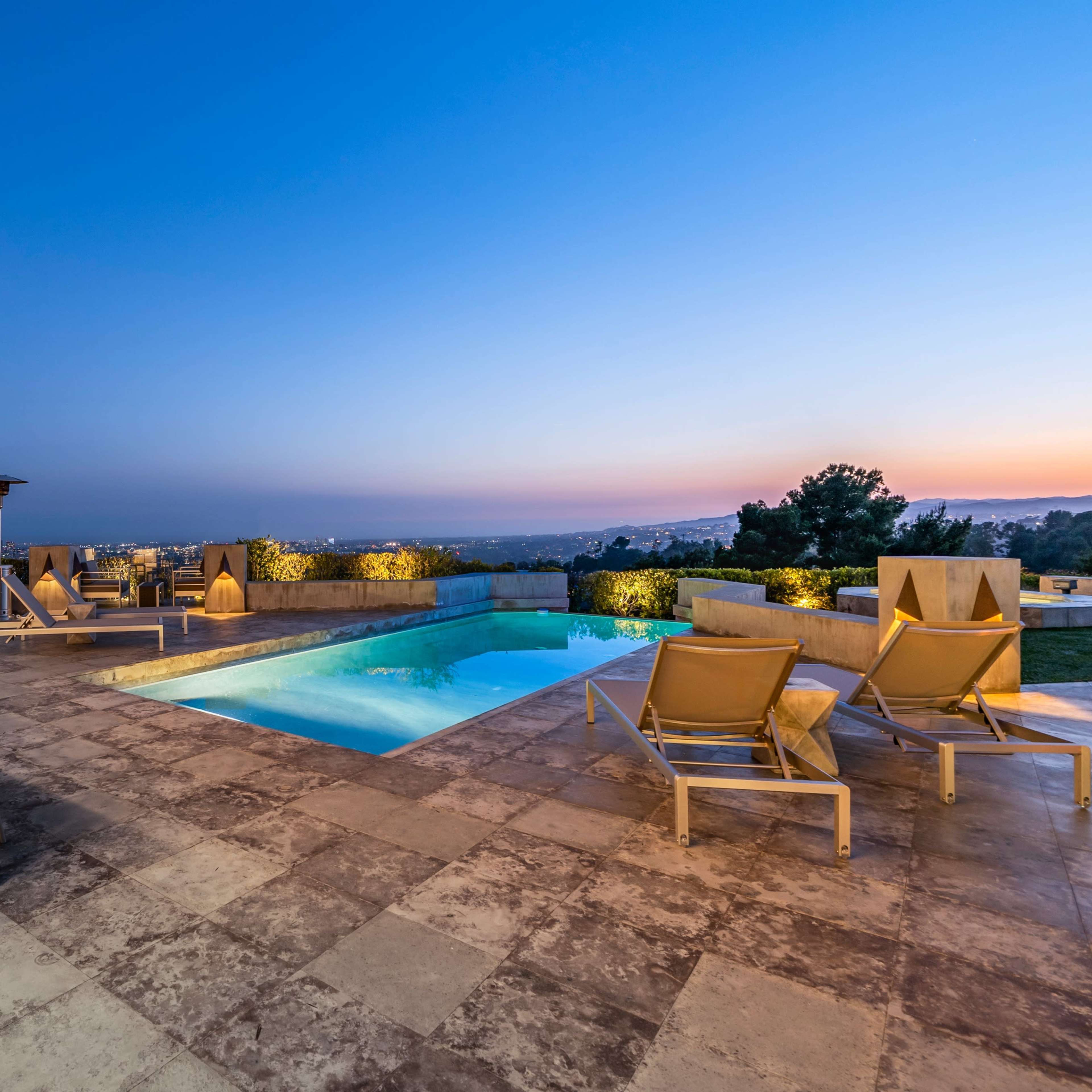 A modern outdoor patio features a swimming pool, lounge chairs, a dining table, and a view of the city skyline at dusk.
