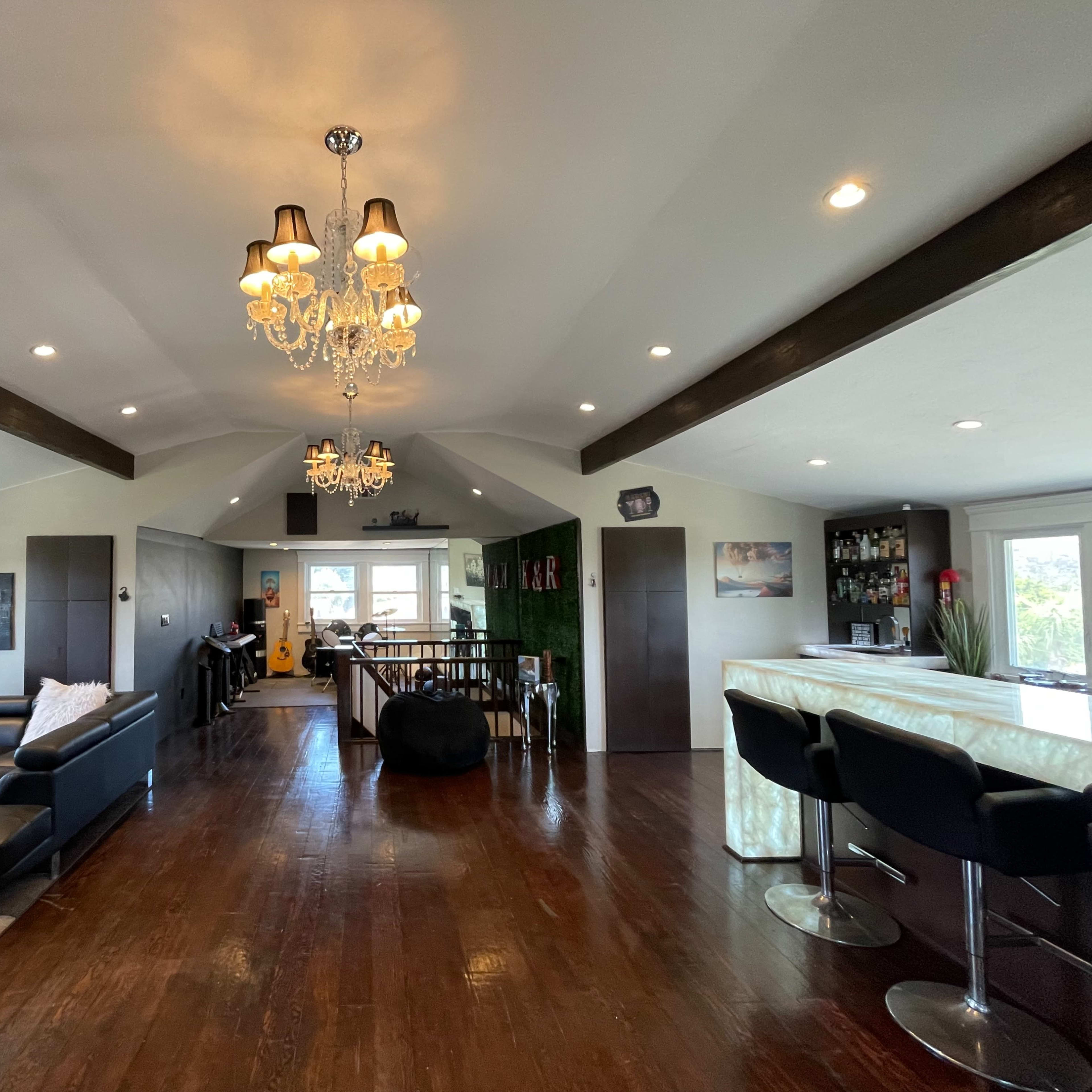 The room features an open living area with a sectional sofa, a bar area with stools, and decorative chandeliers, all set against dark wood flooring and a high ceiling.