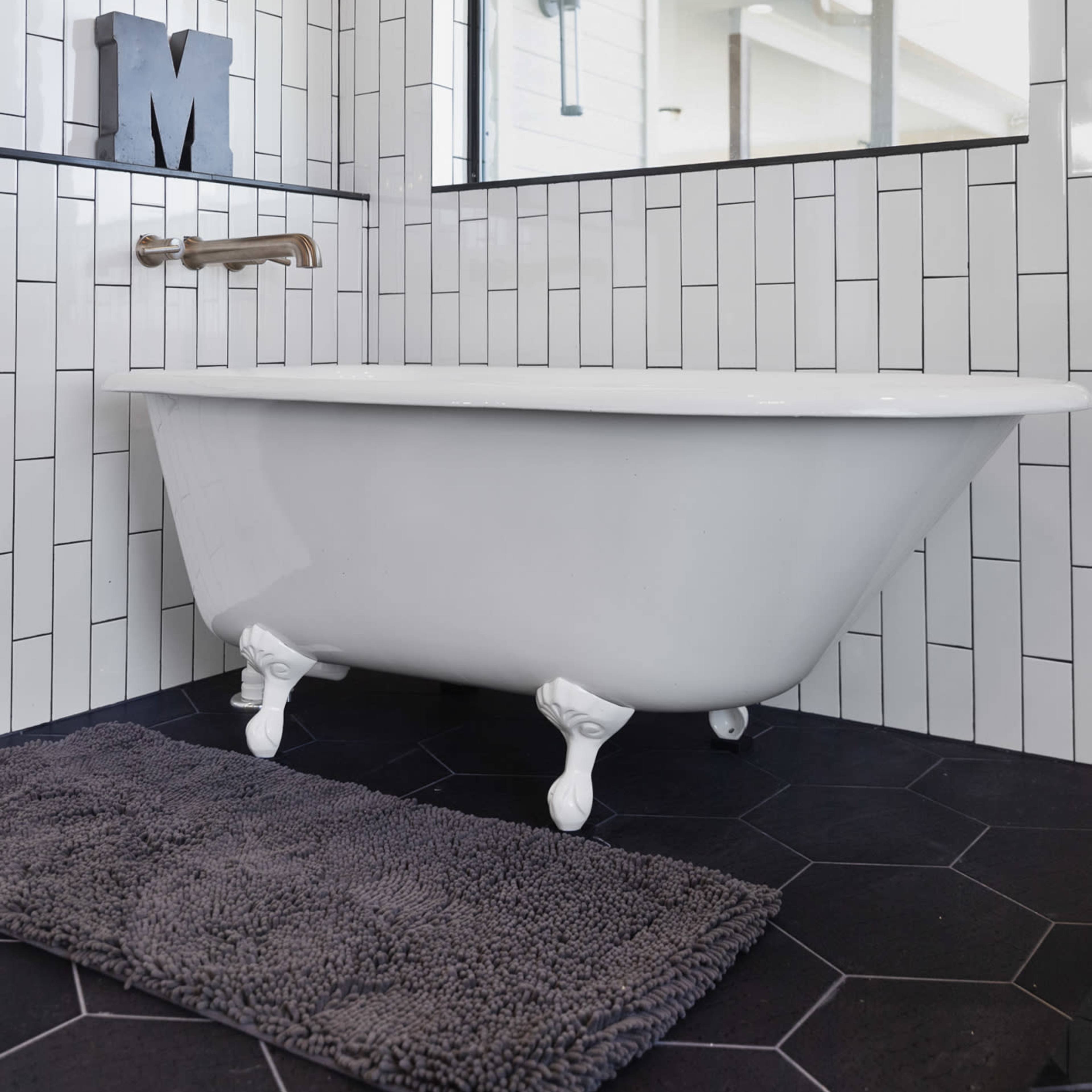 A freestanding white bathtub with claw feet sits on a black tiled floor next to a window in a bathroom with white subway tile walls.