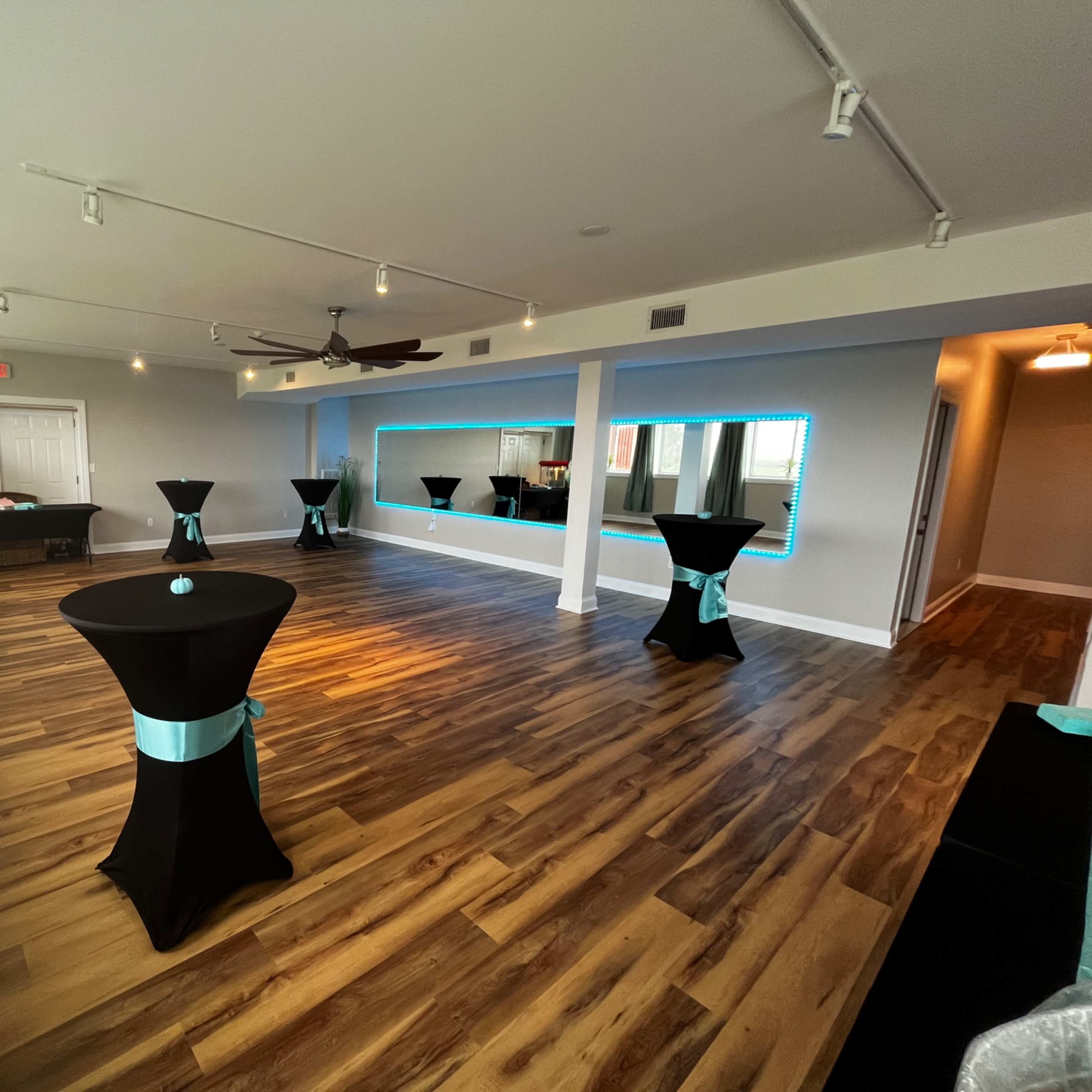The room features several black cocktail tables with turquoise accents arranged across a wooden floor, alongside large windows and a backlit mirror wall.