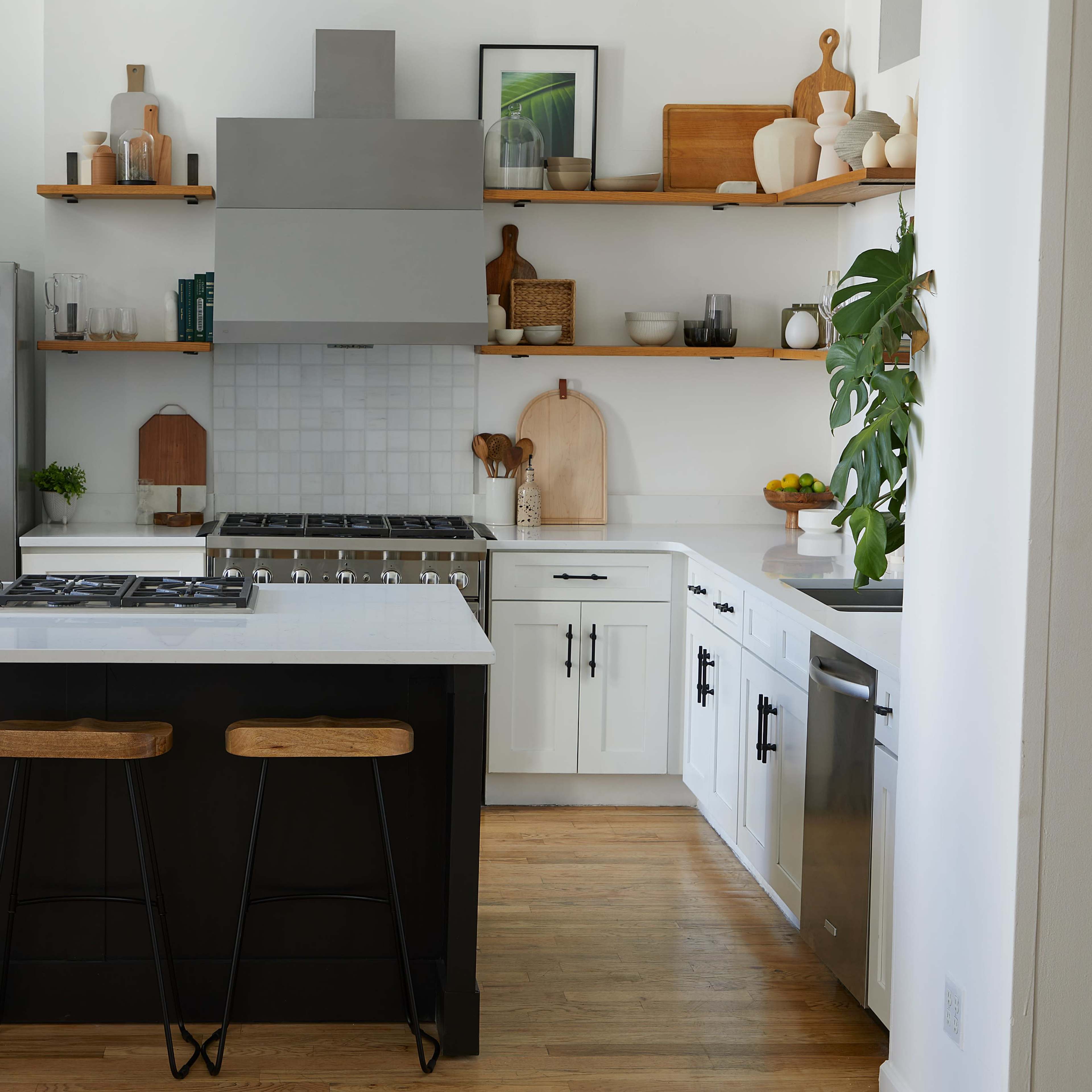 A modern kitchen features a large island with three wooden stools, stainless steel appliances, white cabinets, and open shelving, all set against a light-colored wall.
