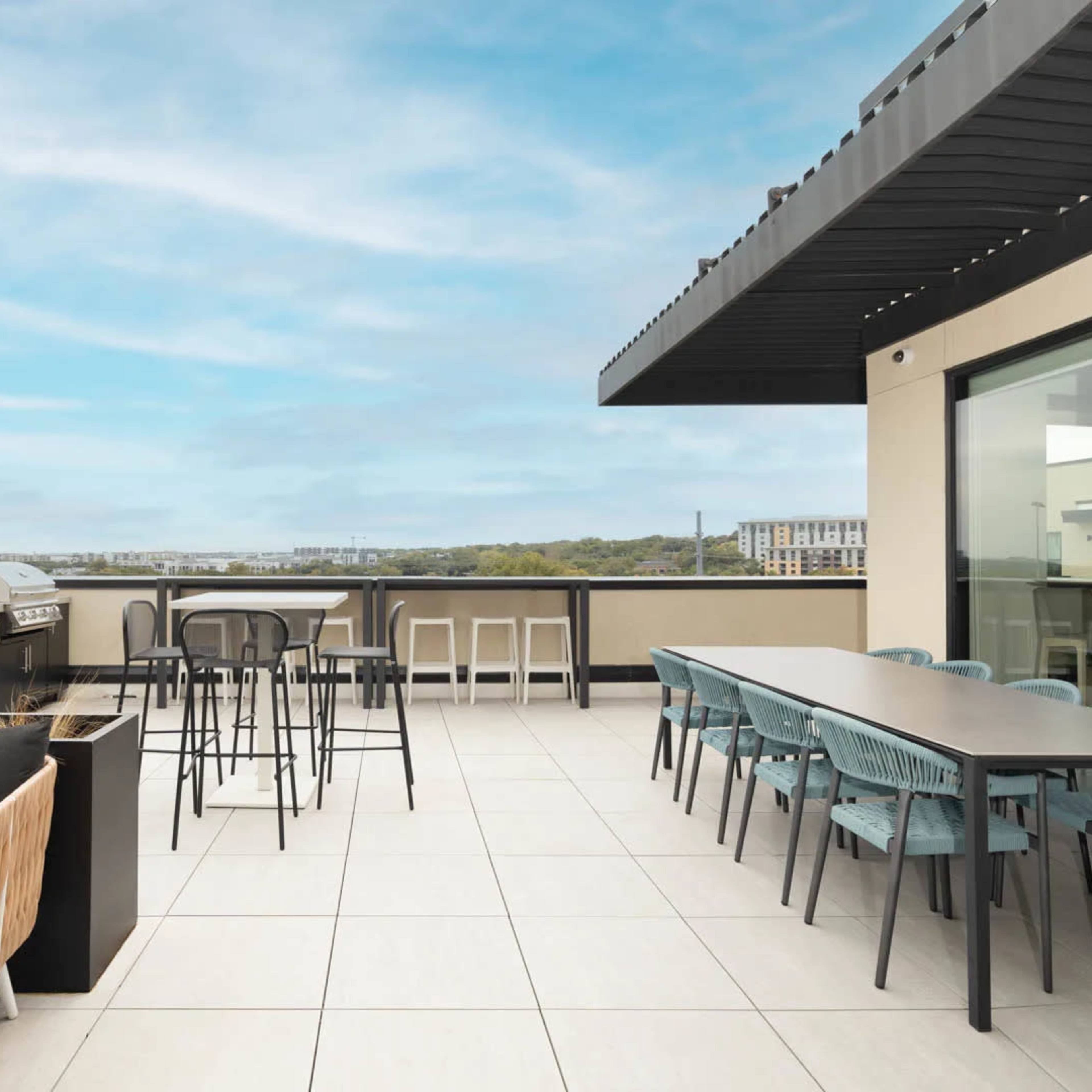 A spacious rooftop terrace featuring seating areas, a grill, and dining tables with a view of the surrounding landscape.