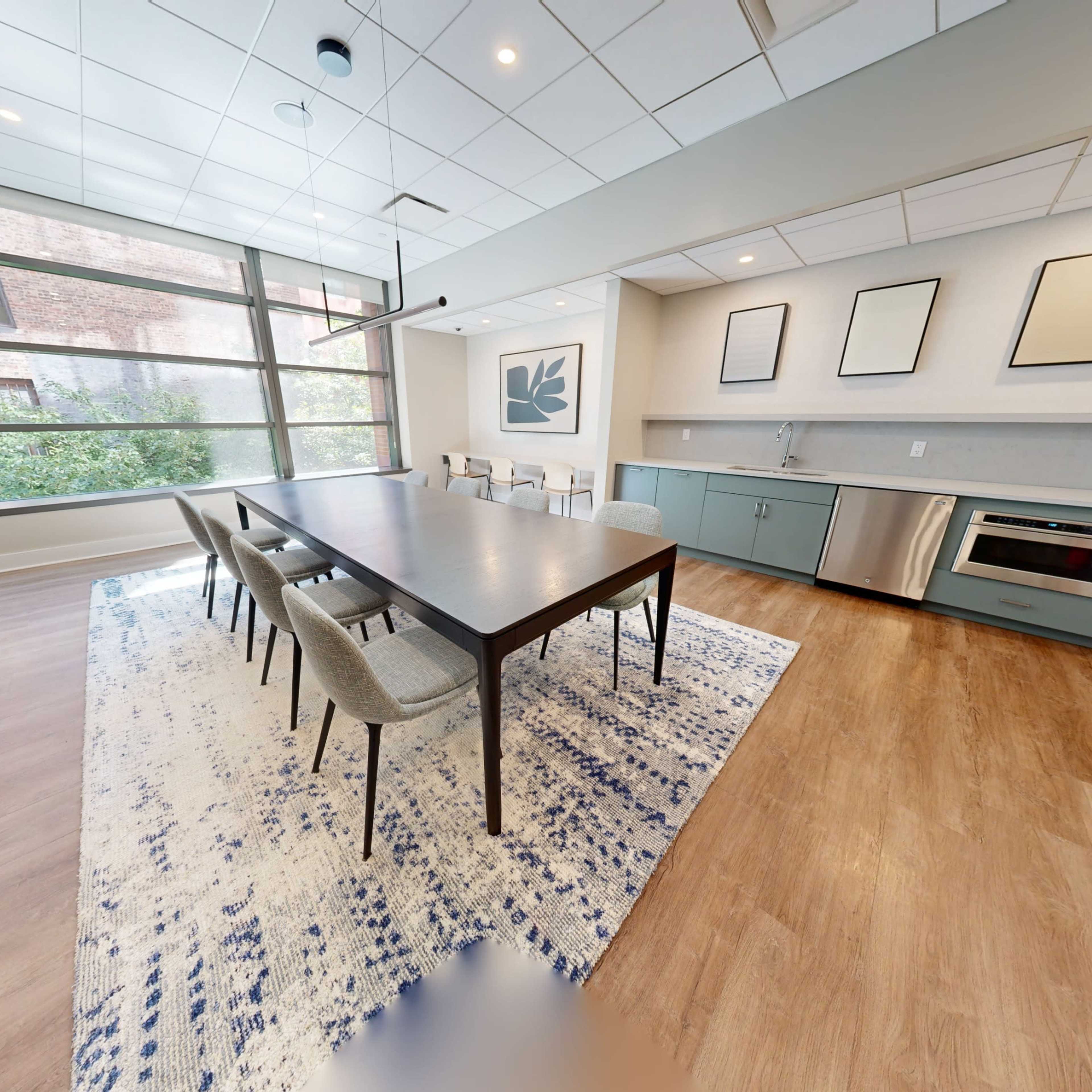 A modern dining area with a large table surrounded by chairs, adjacent to a kitchen space featuring a small oven and light-colored cabinetry, illuminated by natural light through large windows.