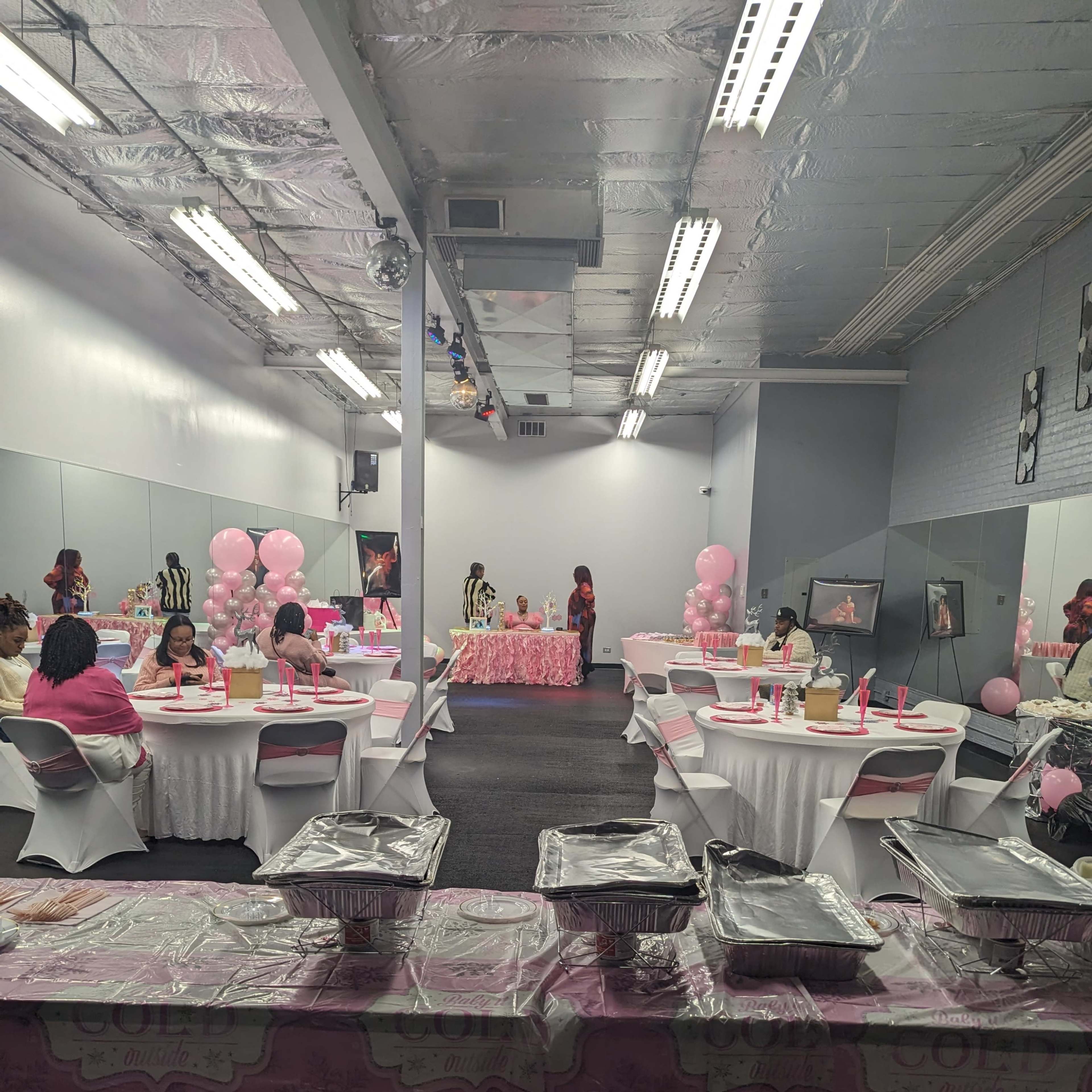 A room is set up for a celebration, featuring tables covered in pink tablecloths, covered dishes, and pink balloons arranged decoratively.