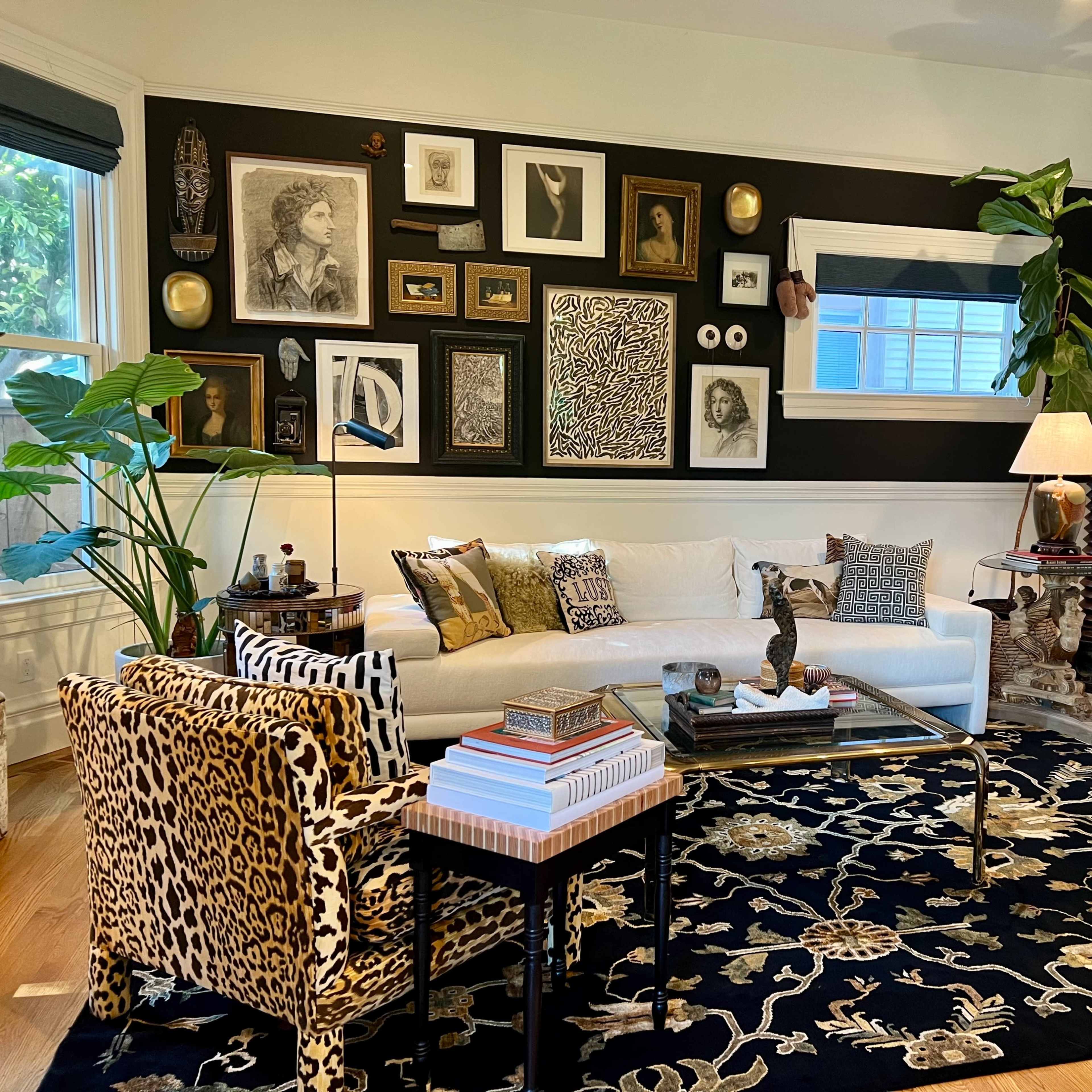 A living room adorned with an array of framed artwork on a dark wall, a white sofa, two leopard print chairs, a glass coffee table, and indoor plants.