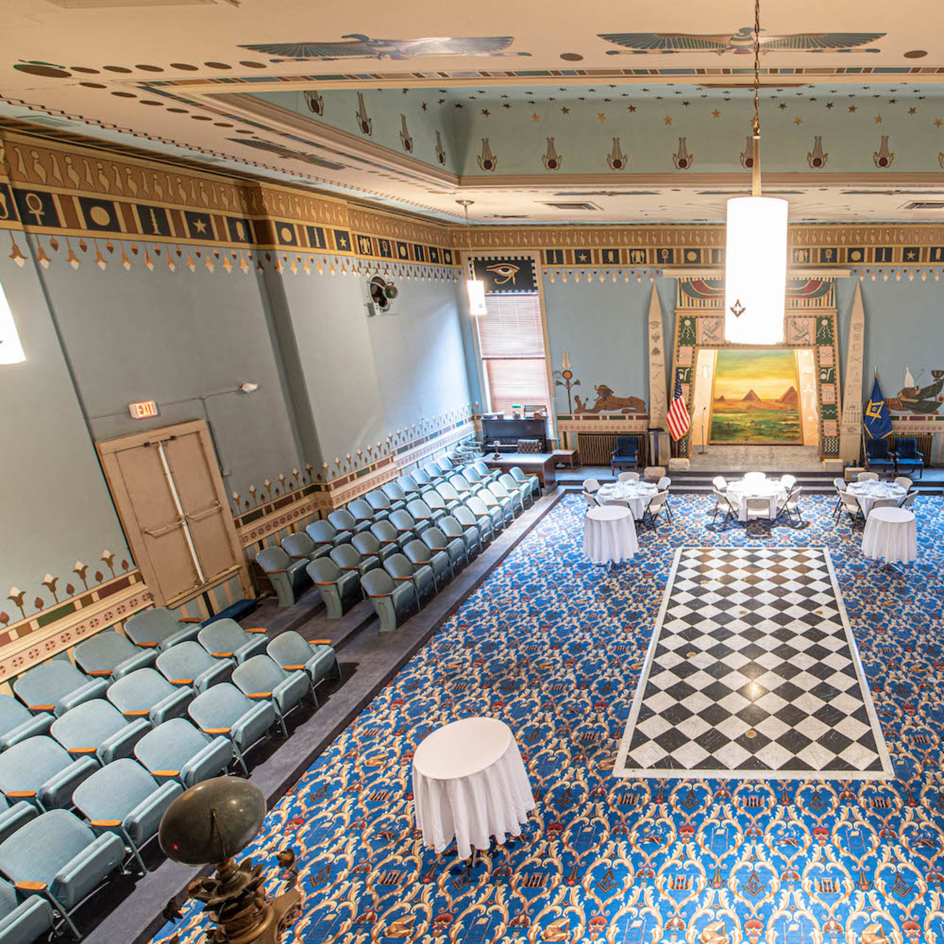 A spacious room set up for an event, featuring a checkered floor, seated rows of chairs, and decorative walls with murals.